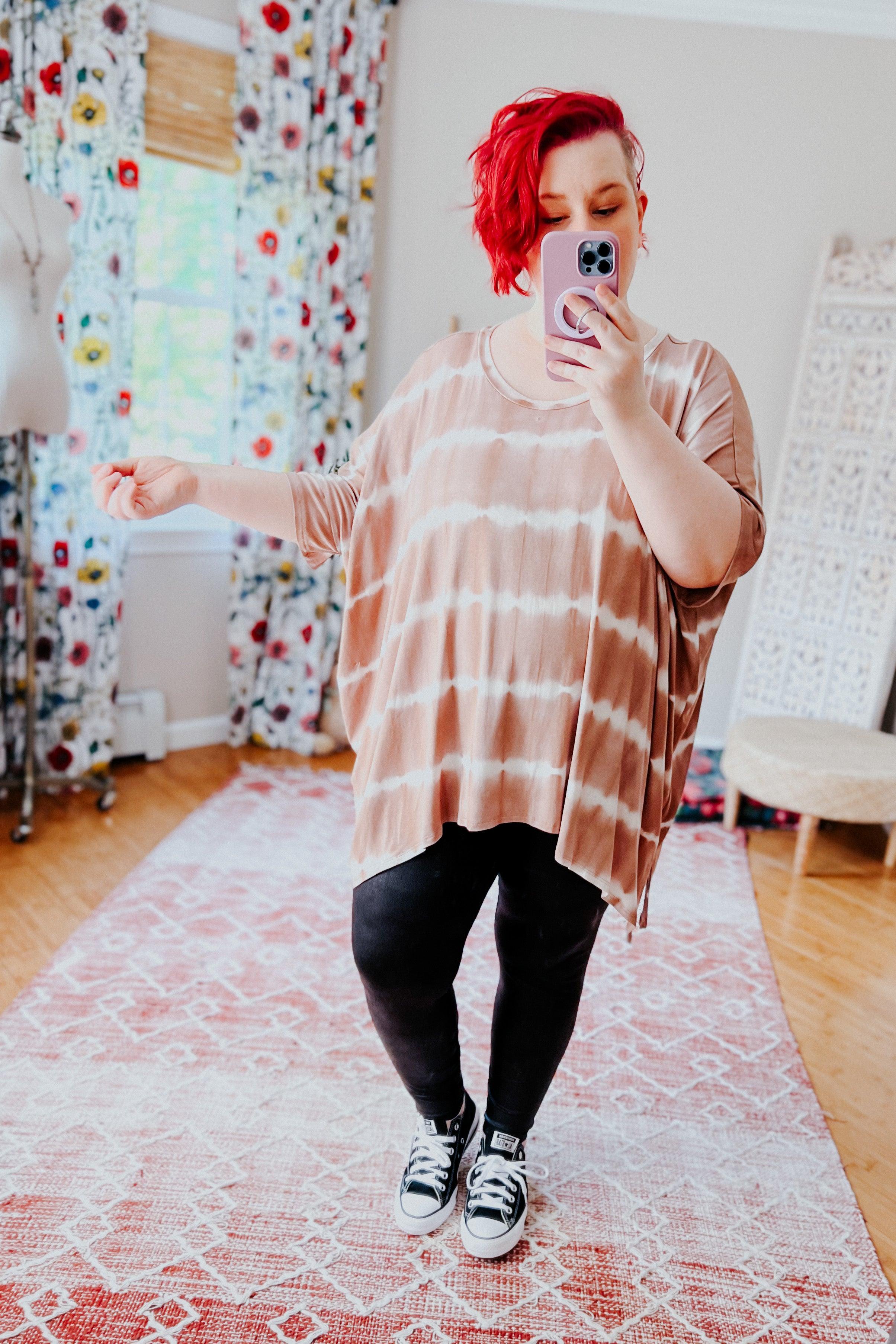 Woman with red hair wearing a loose tan and white striped tie dye tunic and black pants taking a mirror selfie indoors