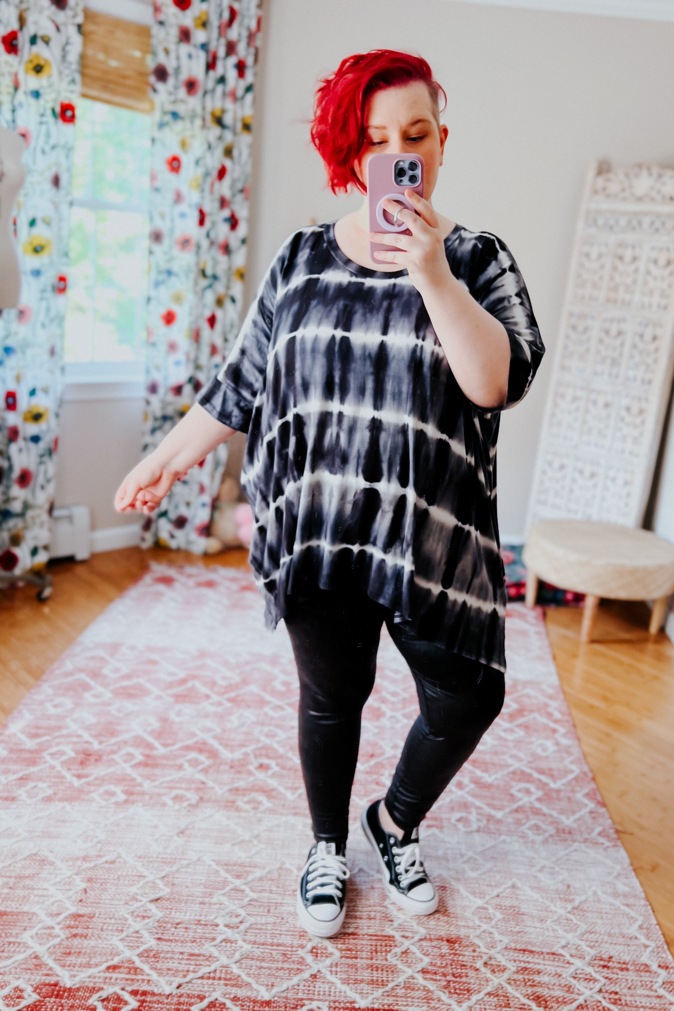 Woman with bright red hair wearing a black striped tie dye tunic and black leggings posing in a colorful room