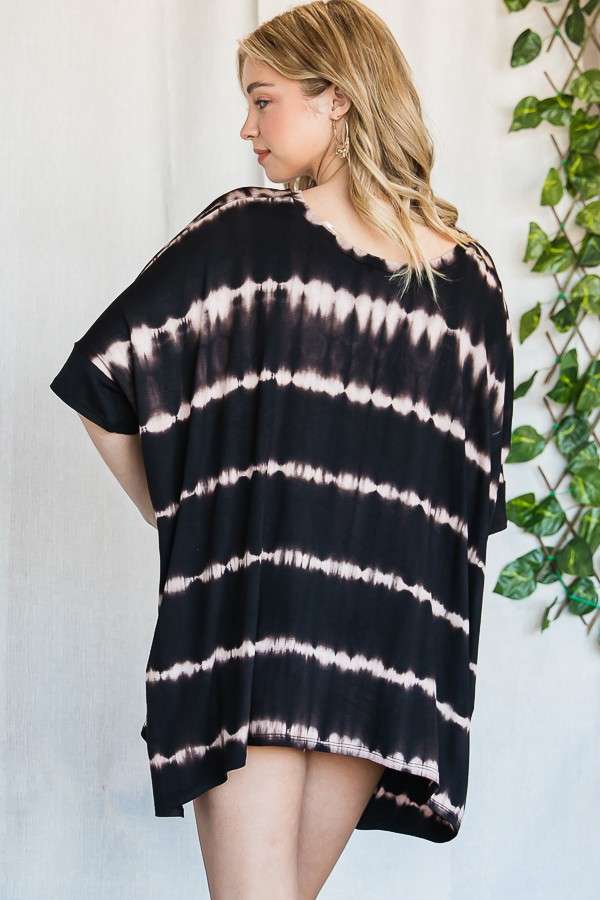 Woman wearing a loose black and white striped tie dye tunic with short sleeves standing against a light wall with greenery nearby