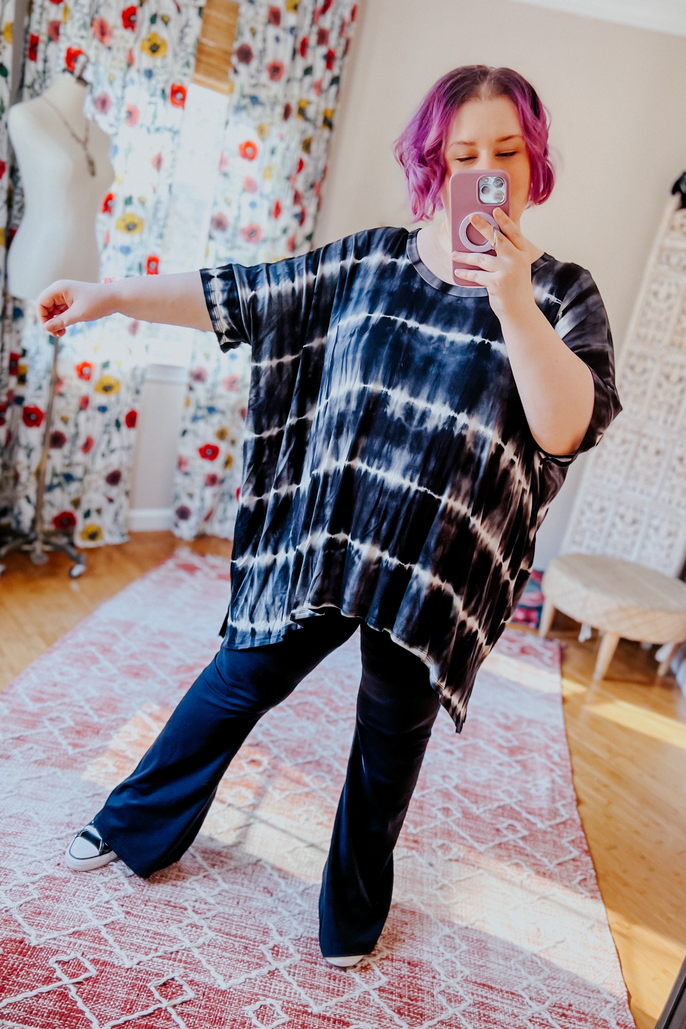 Woman taking a mirror selfie wearing a loose black striped tie dye tunic and black pants in a bright room with floral curtains