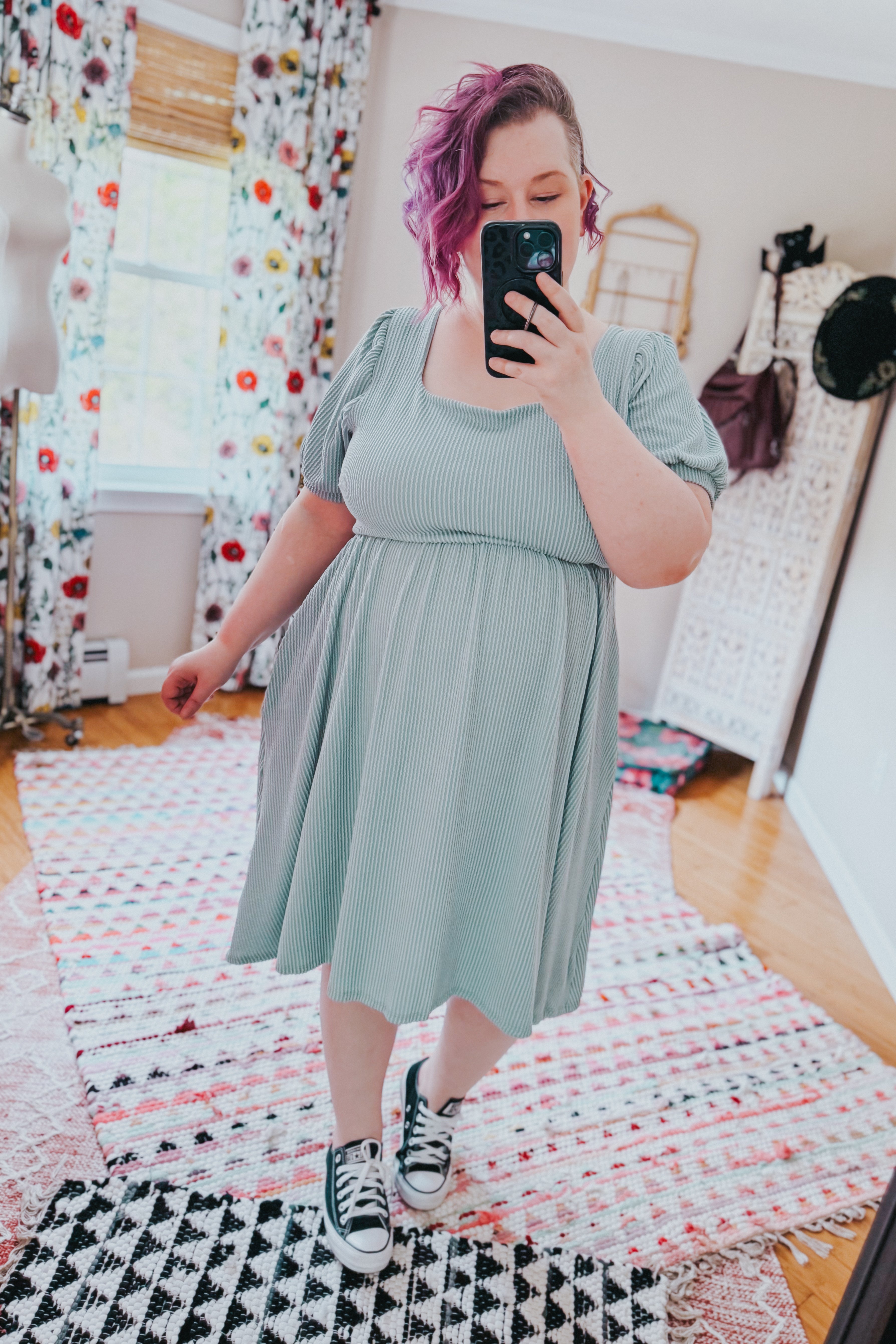 Plus size model wearing a light sage cable ribbed knit dress and black sneakers in a bright room with floral curtains