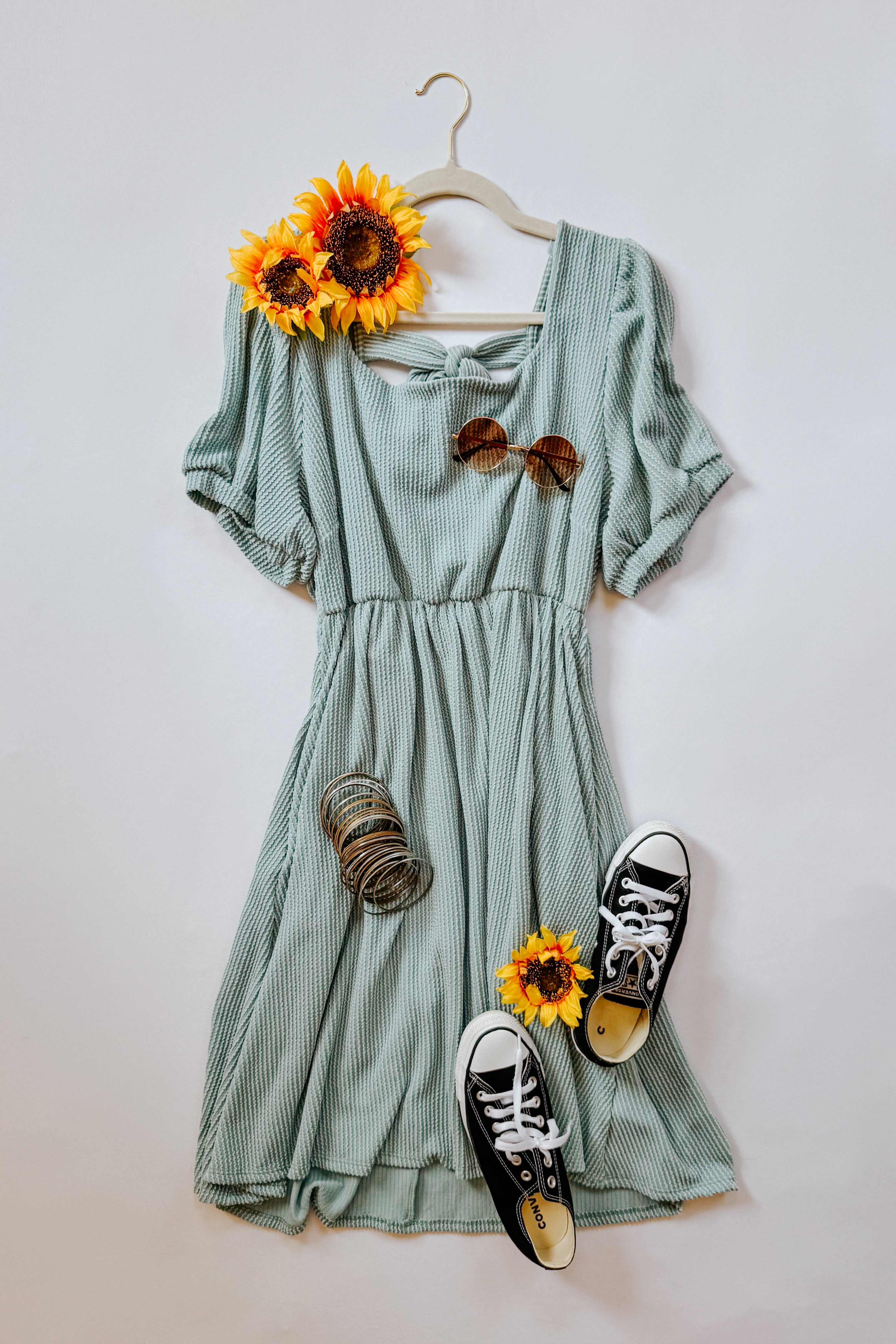 Light sage cable ribbed knit dress styled with round sunglasses, black sneakers, bangles, and sunflower accents on a hanger
