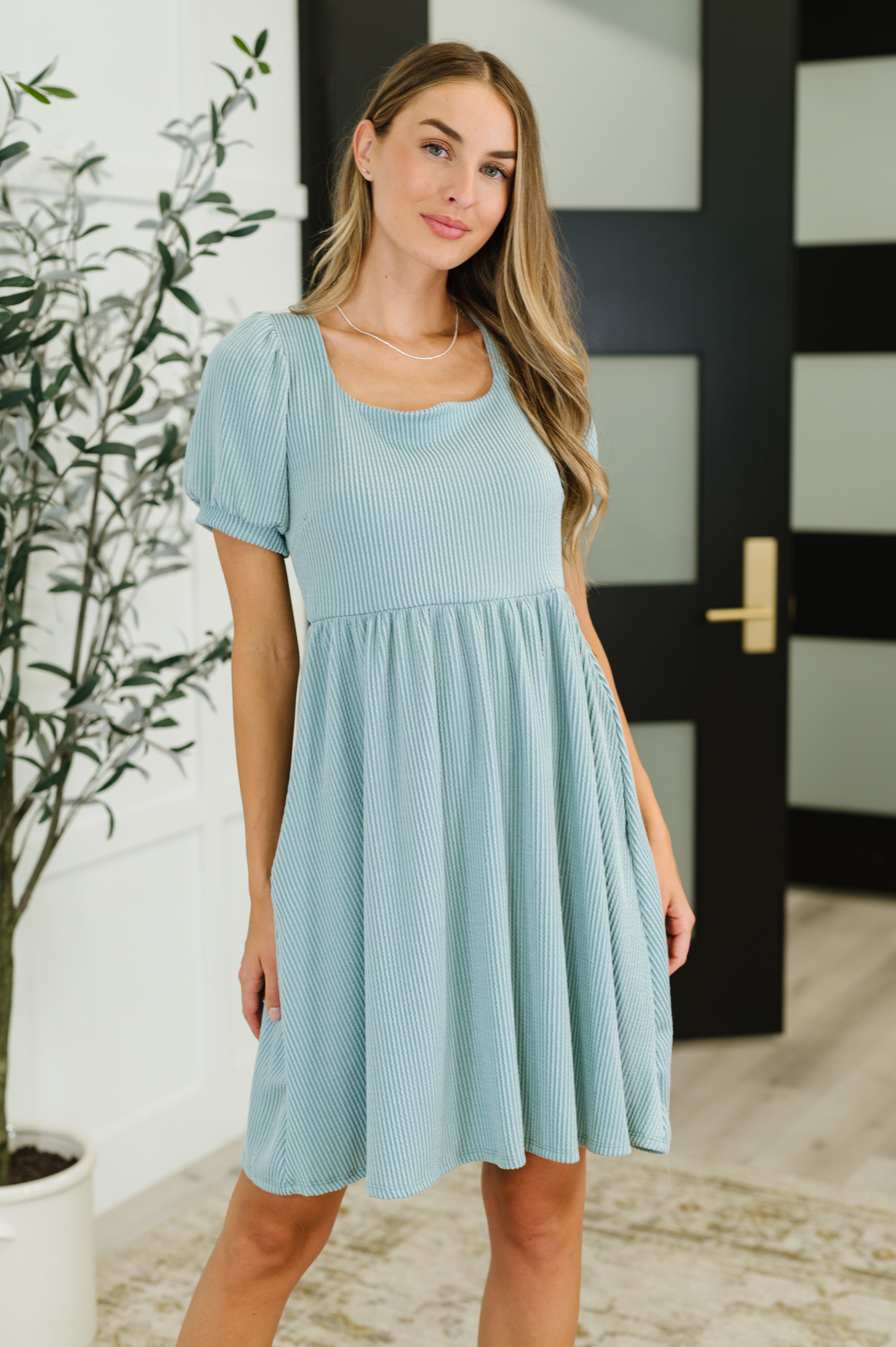 Woman wearing a light sage cable ribbed knit dress with puff sleeves standing indoors near a potted plant and black door