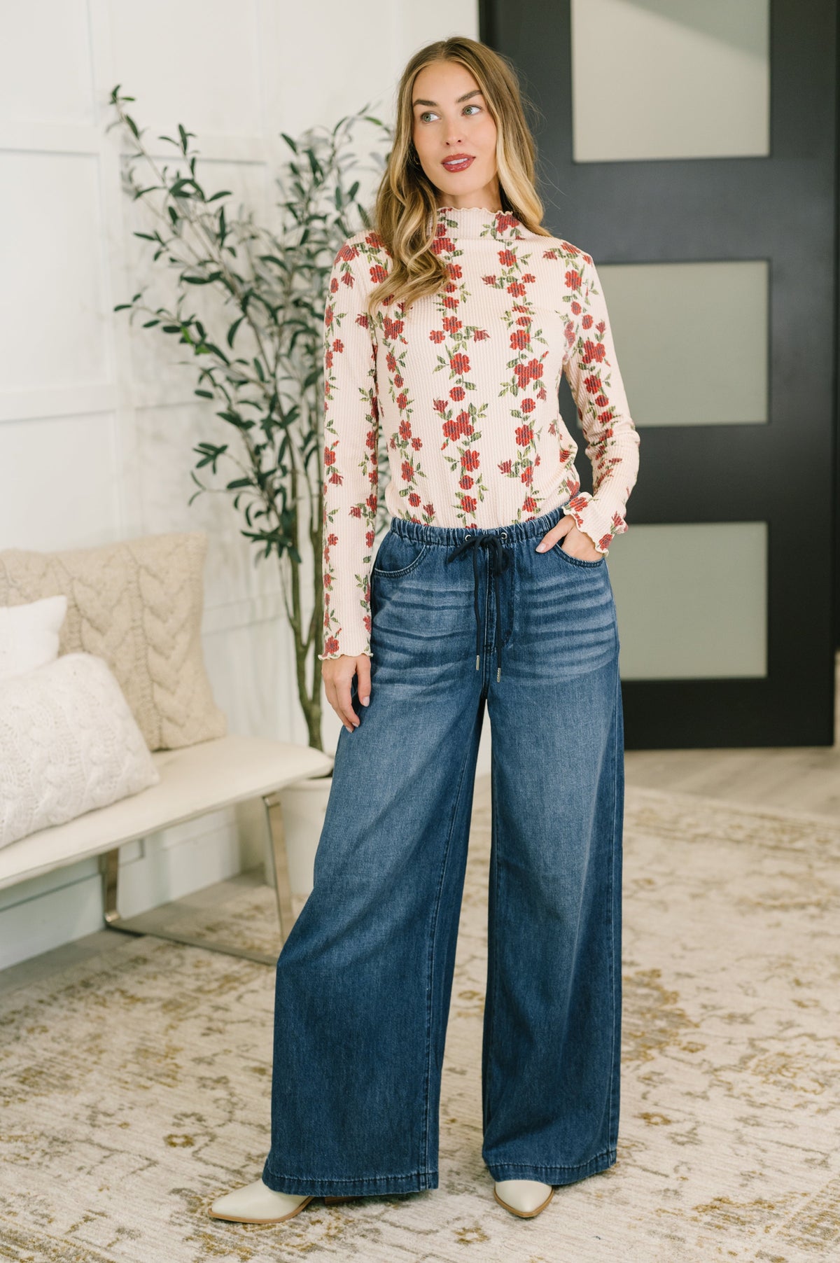 Woman wearing a beige high neck floral ribbed top with long sleeves and wide leg jeans in a bright modern living room