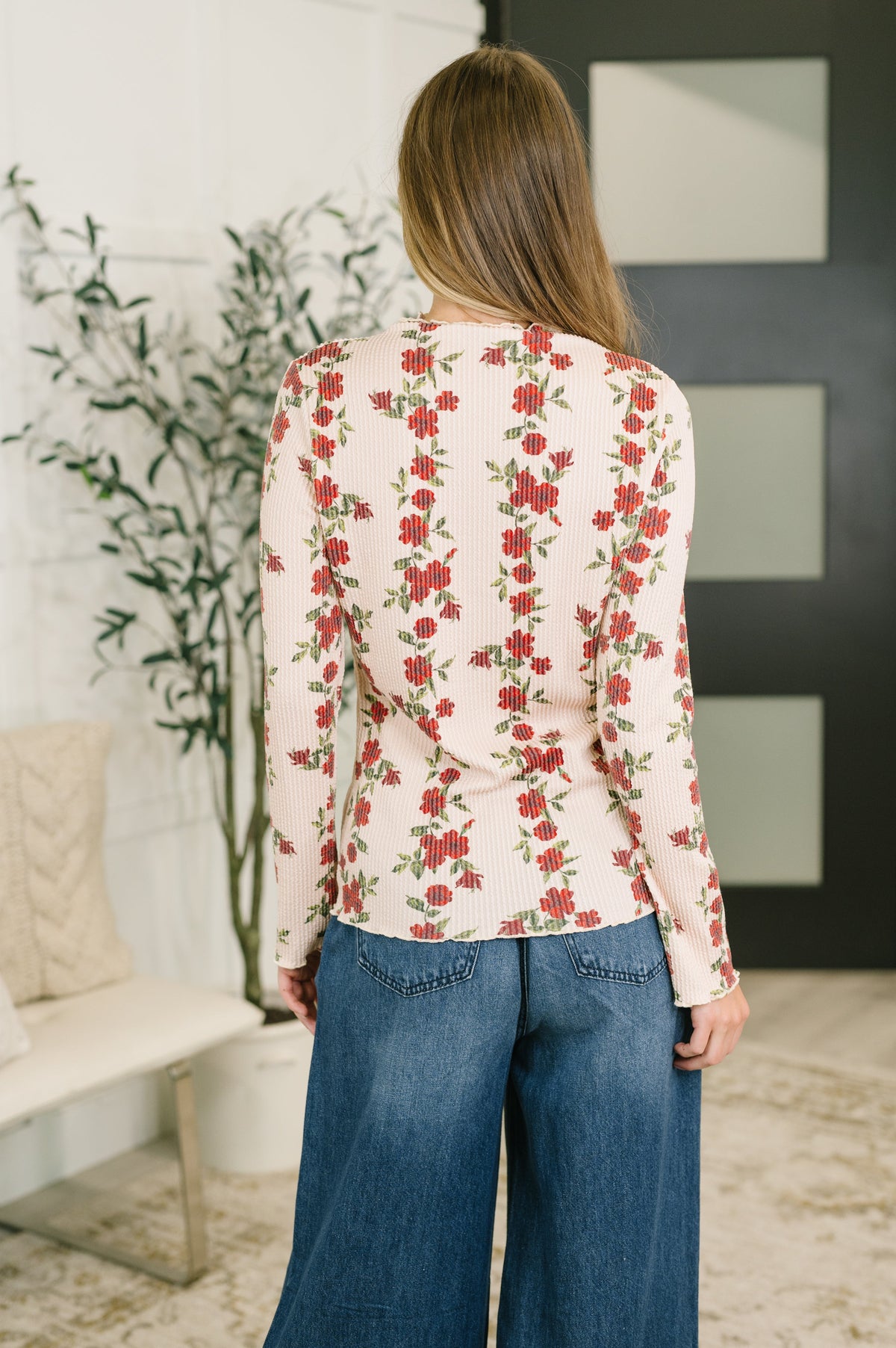 Back view of a woman in a beige floral cable ribbed high neck top with long sleeves and wide leg jeans in a bright modern interior