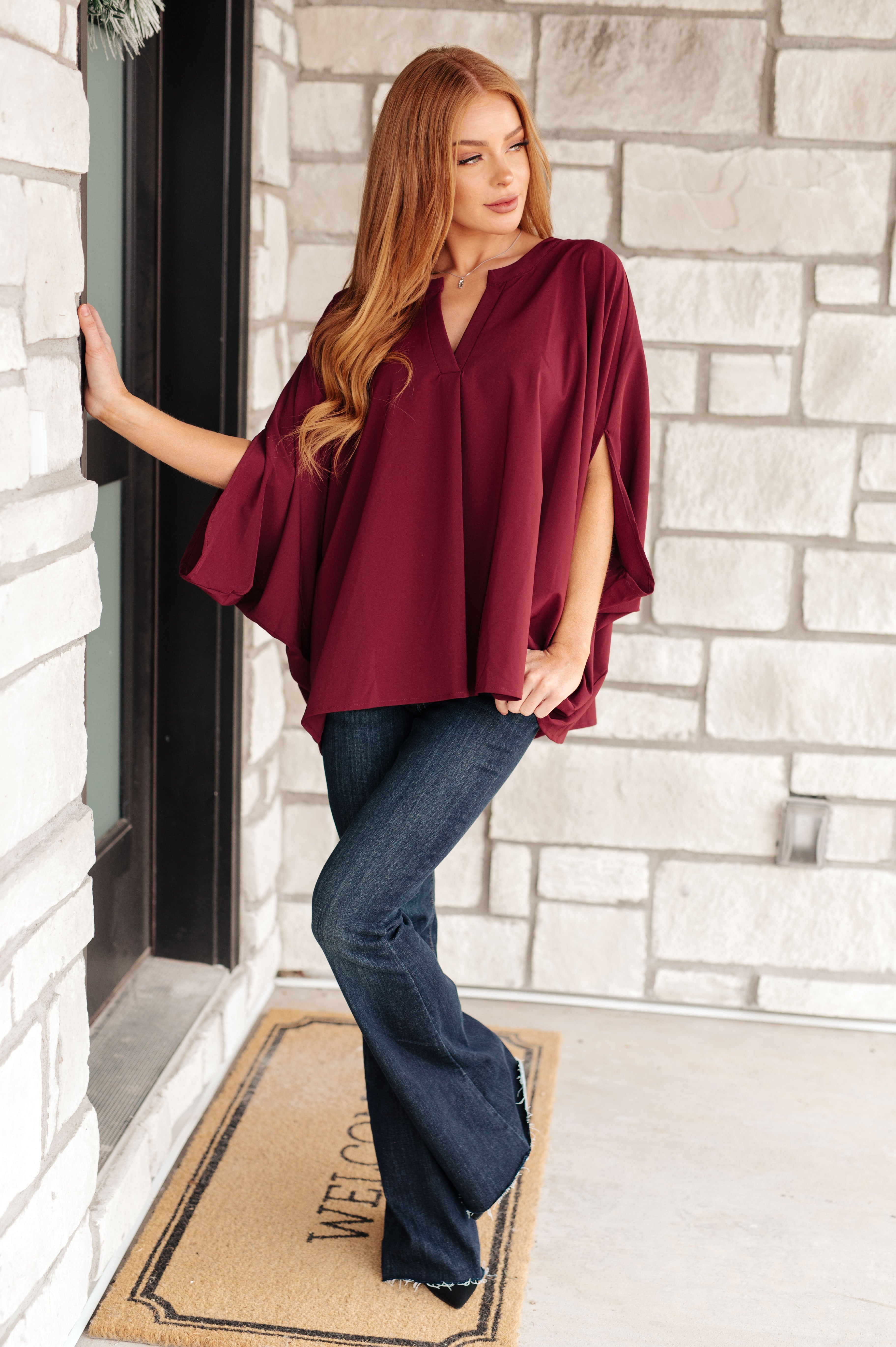 Woman wearing a loose wine-colored blouse with split sleeves paired with dark flared jeans standing by a stone wall