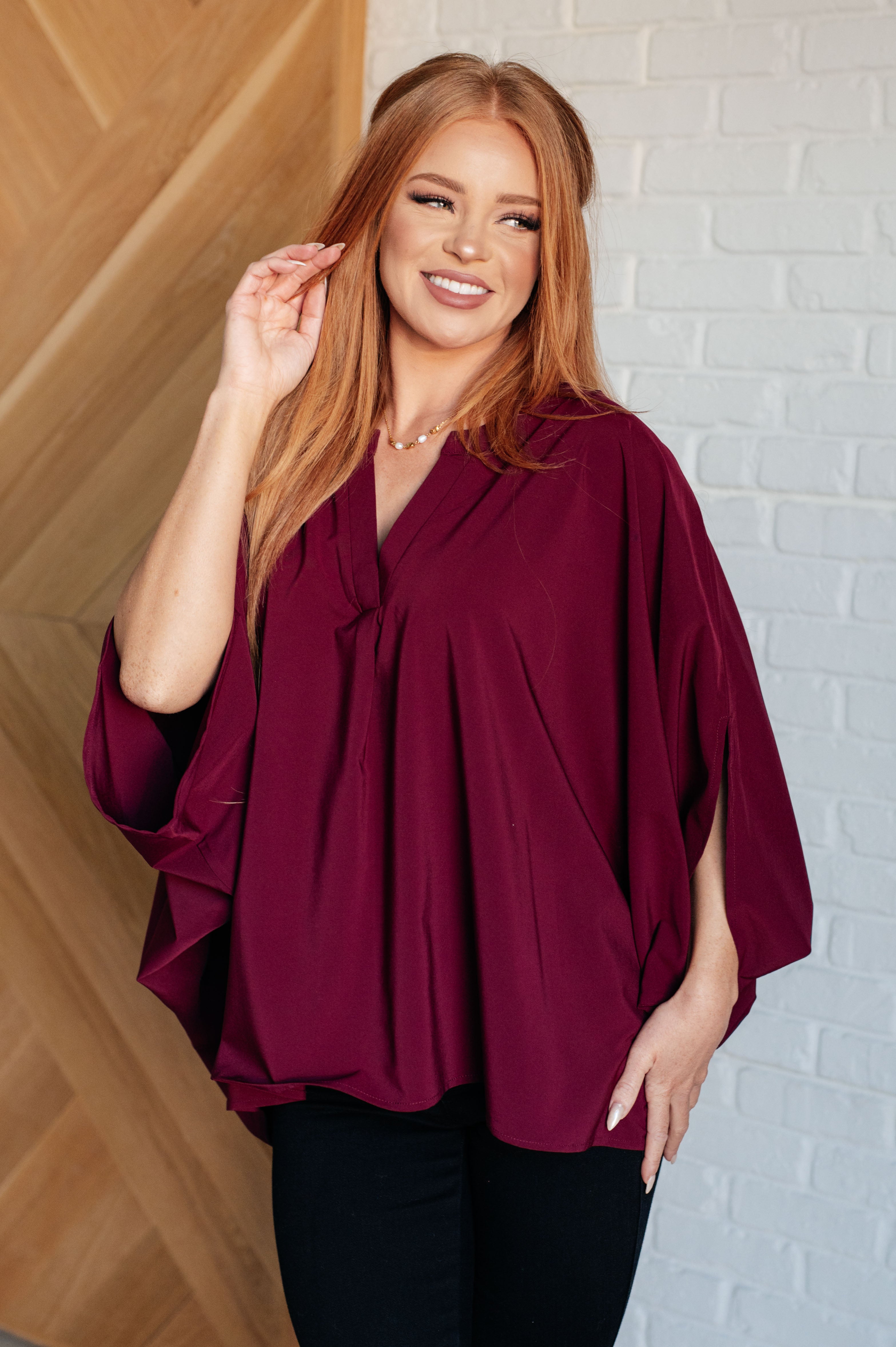 Woman wearing a wine-colored loose blouse with draped sleeves and a subtle v-neck, smiling indoors by a wood wall