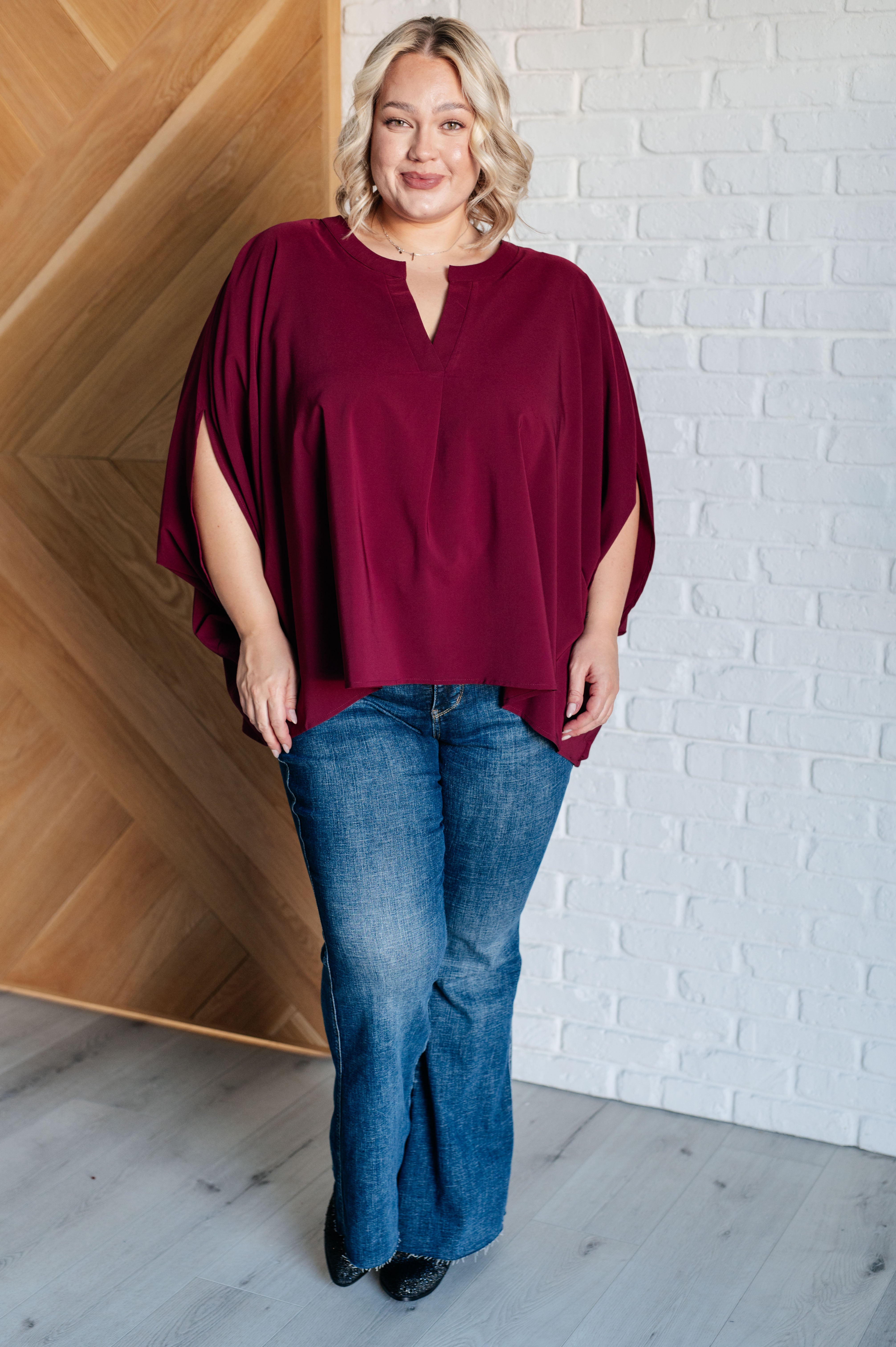 Plus size model wearing a wine-colored loose blouse with split sleeves paired with blue jeans and black shoes indoors