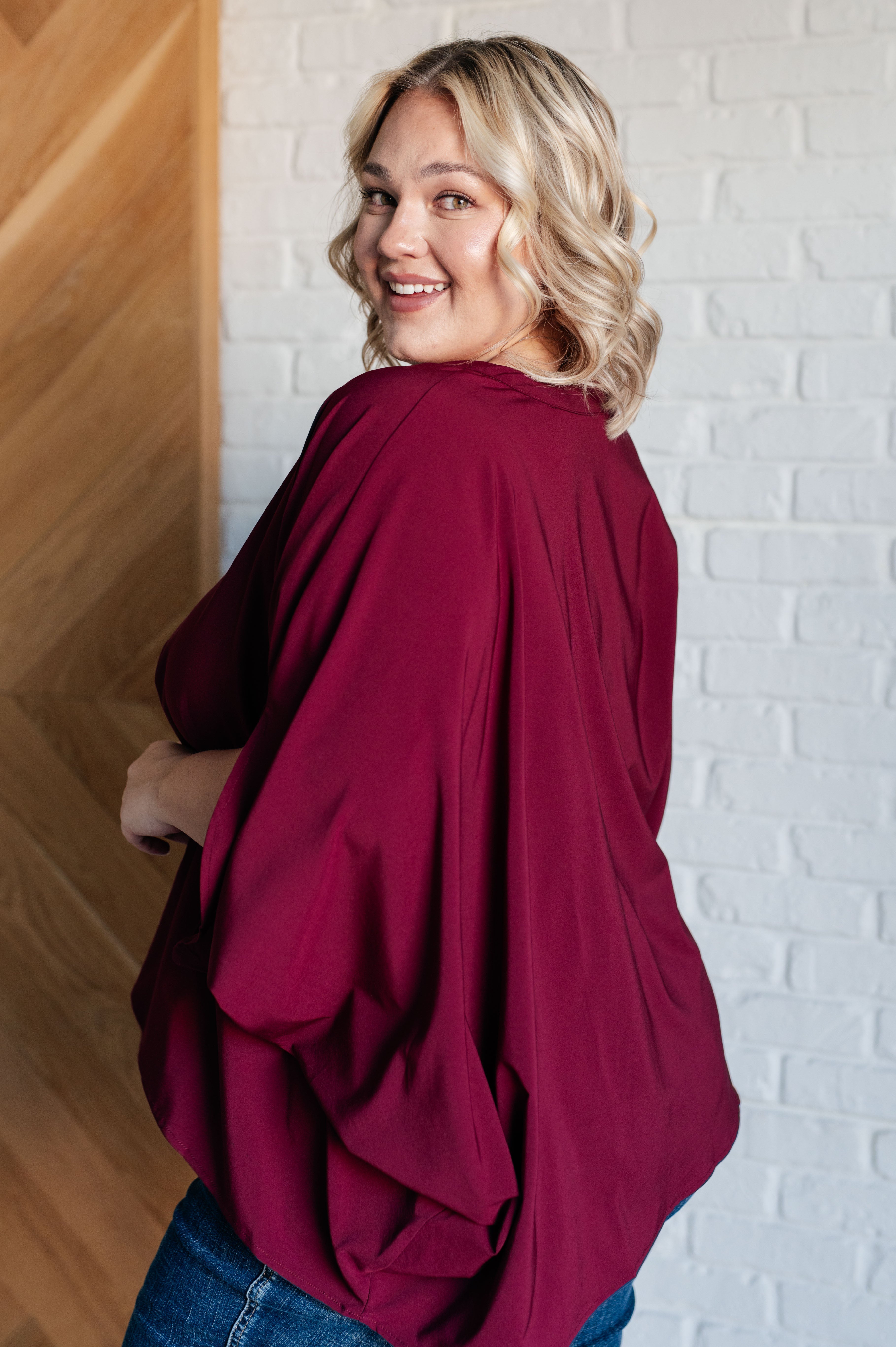 Smiling woman wearing a loose-fitting wine-colored blouse with draped sleeves standing indoors by a wooden and brick wall