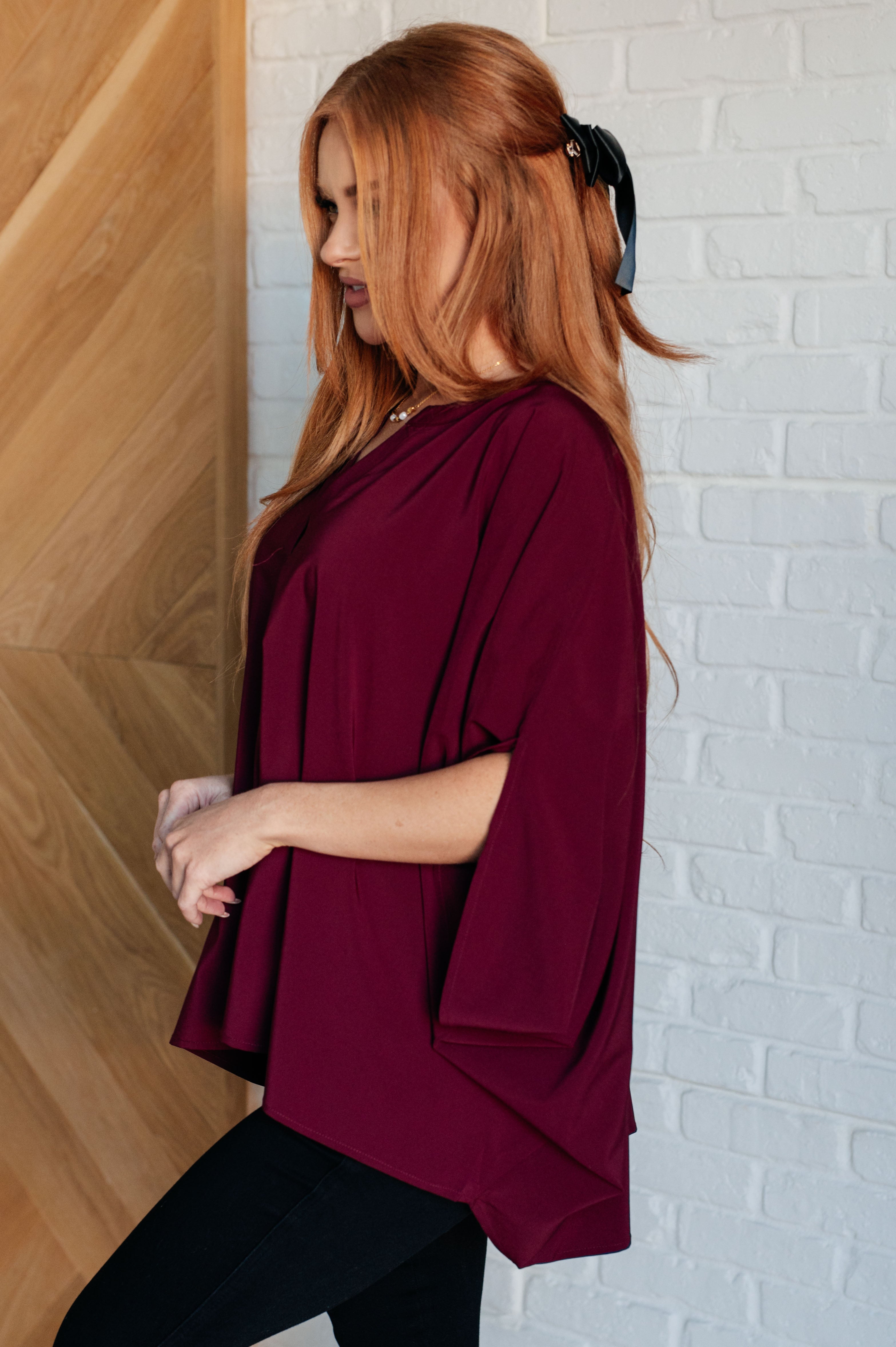 Woman with red hair wearing a loose wine-colored blouse with wide sleeves and black pants standing by white brick wall