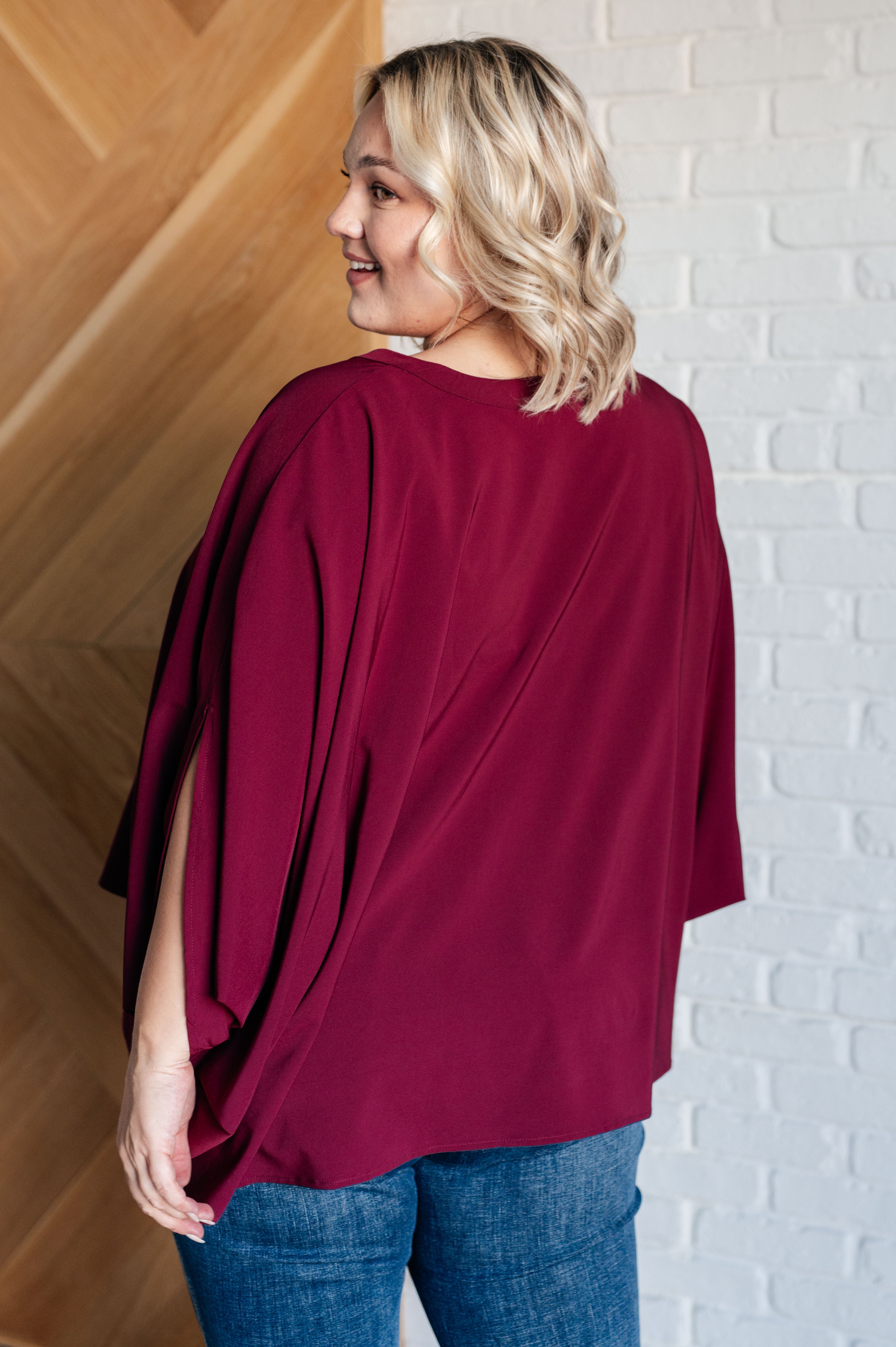 Woman wearing a wine-colored loose blouse with open slit sleeves paired with blue jeans in a bright indoor setting