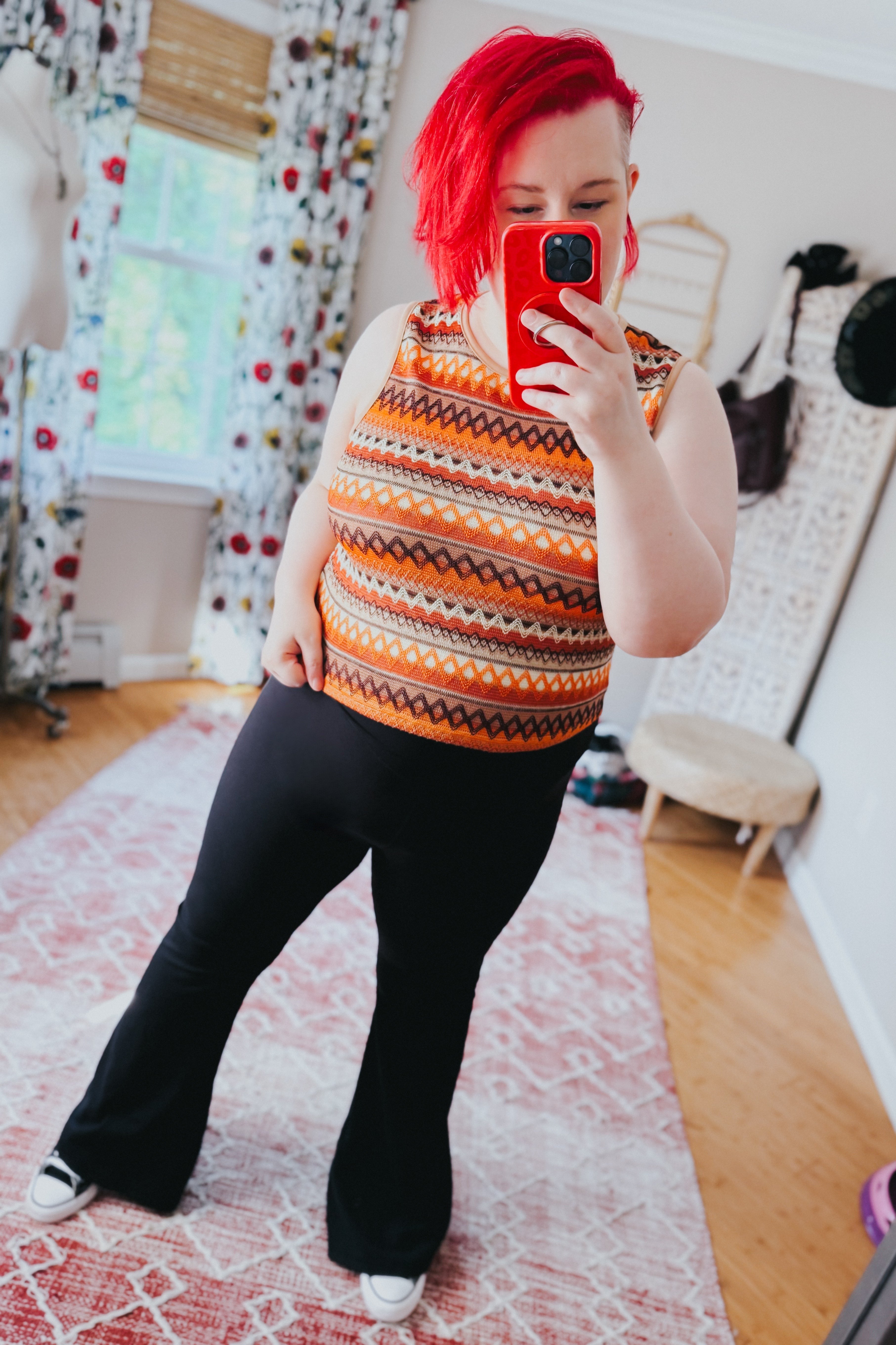 Woman with bright red hair wearing an orange patterned knit tank and black flared pants taking a selfie indoors