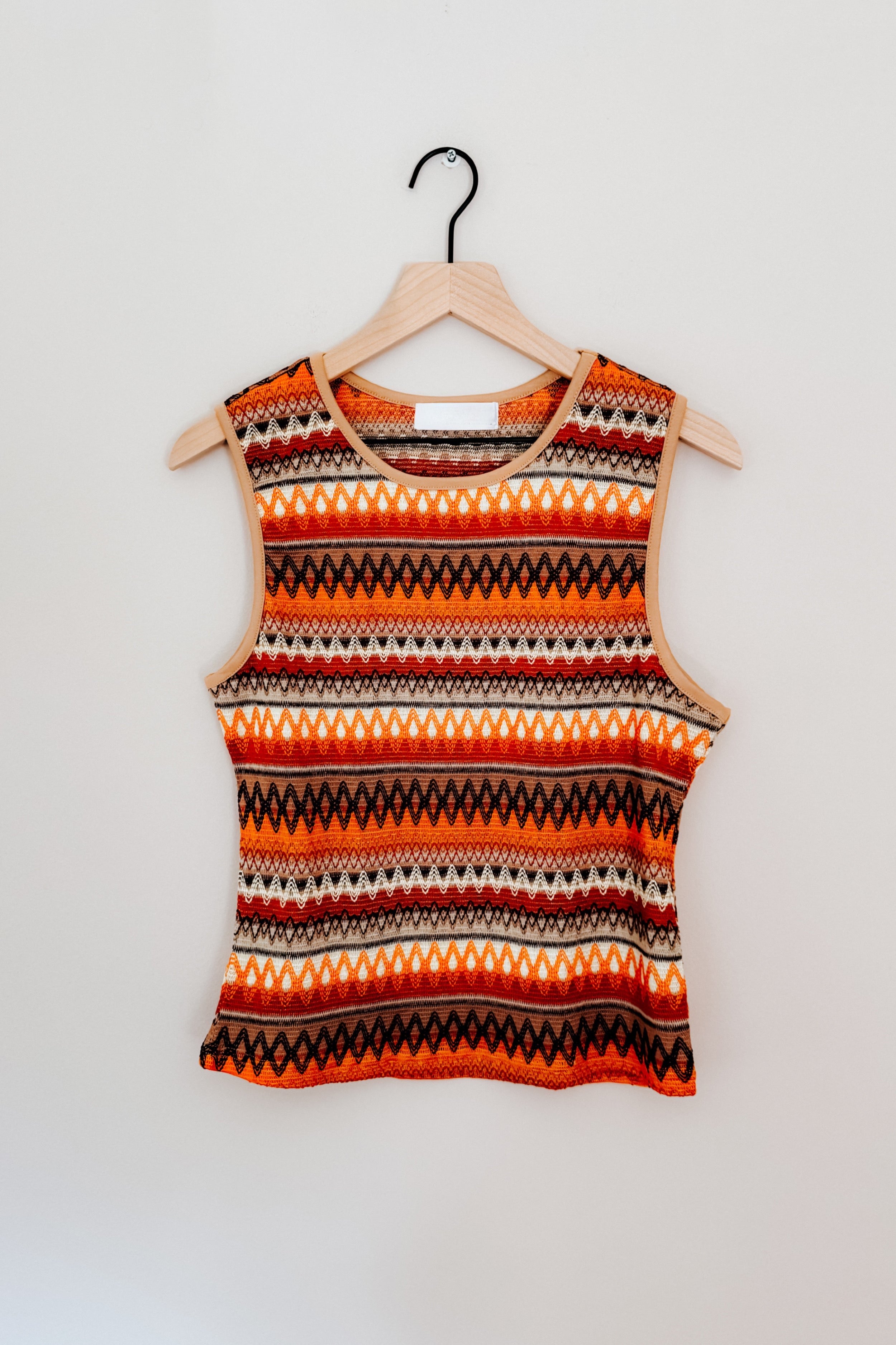 Sleeveless knit tank with bold orange and brown geometric stripes hanging on wooden hanger against white wall