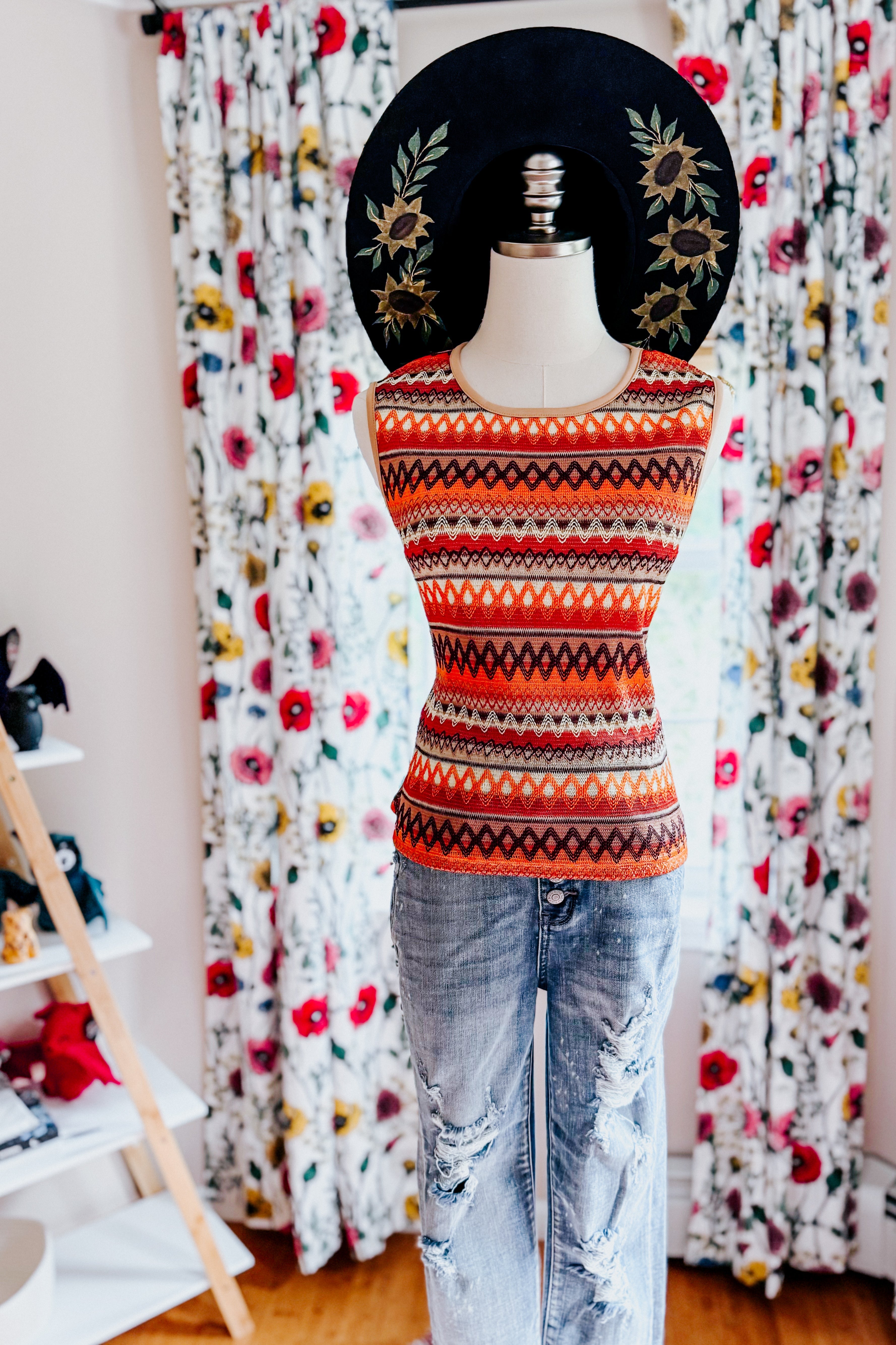 Mannequin dressed in an orange knit tank with geometric patterns paired with distressed light wash jeans and black hat with floral design