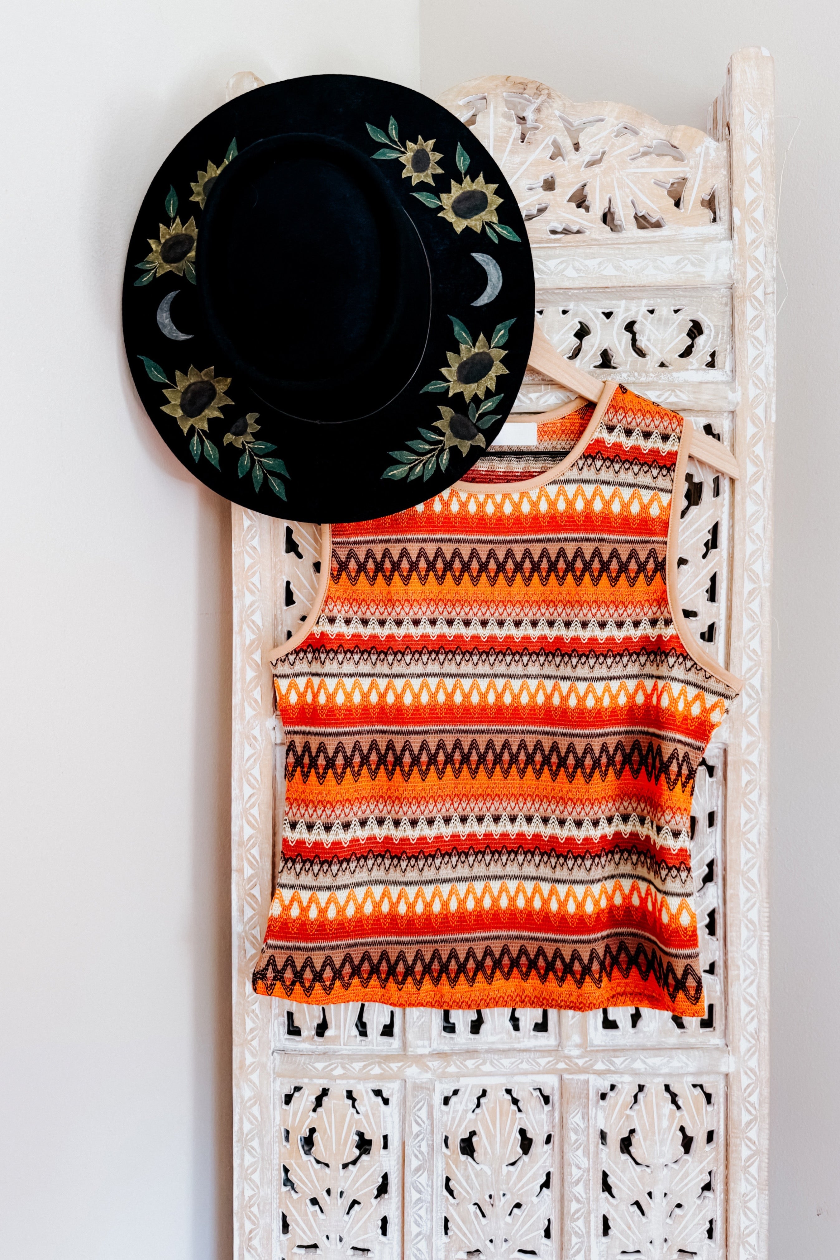 Orange knit tank with geometric stripes hanging on carved white wooden panel beside black hat with sunflower and moon embroidery