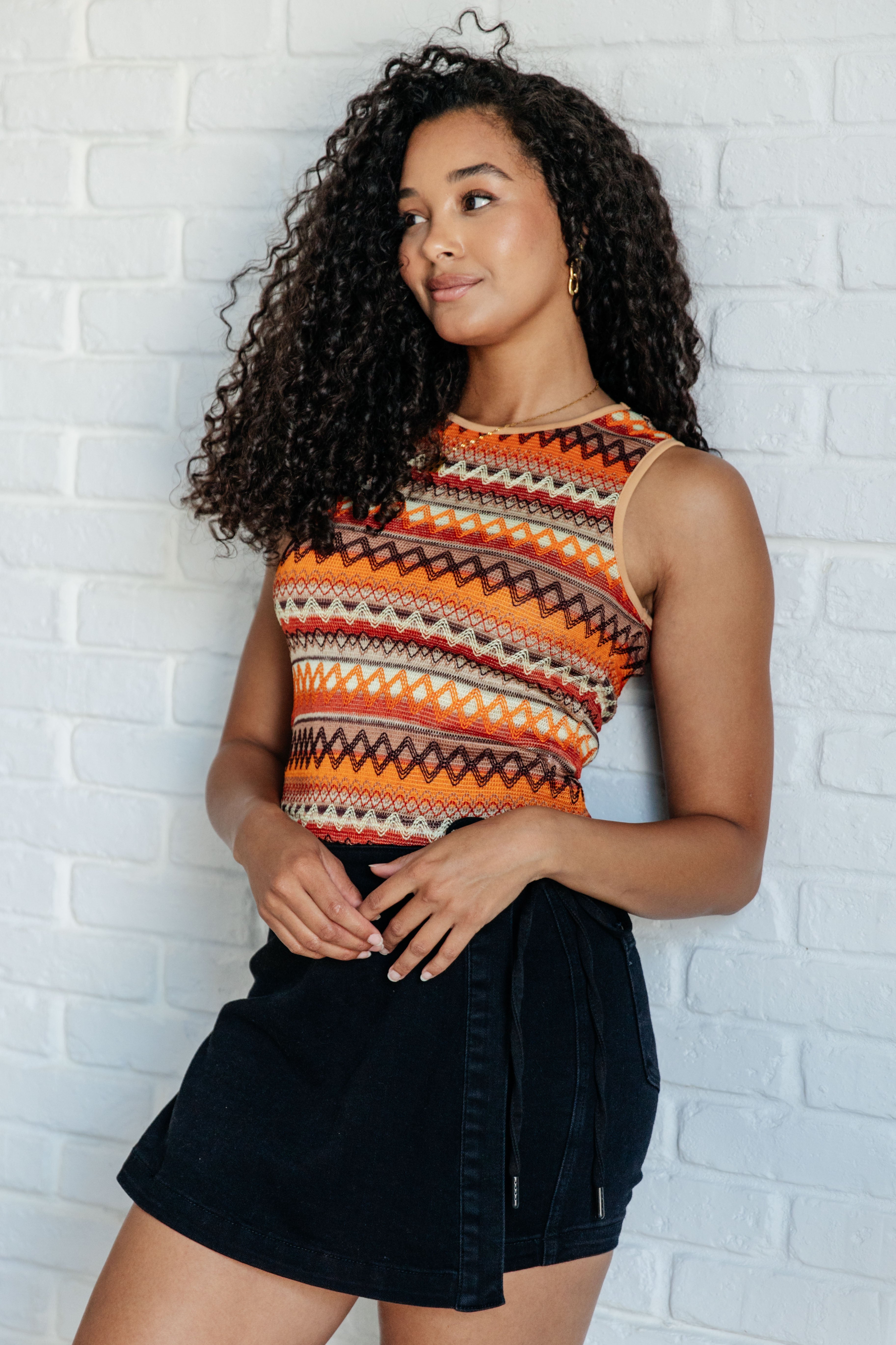Woman wearing an orange knit tank with geometric patterns and a black skirt, standing against a white brick wall