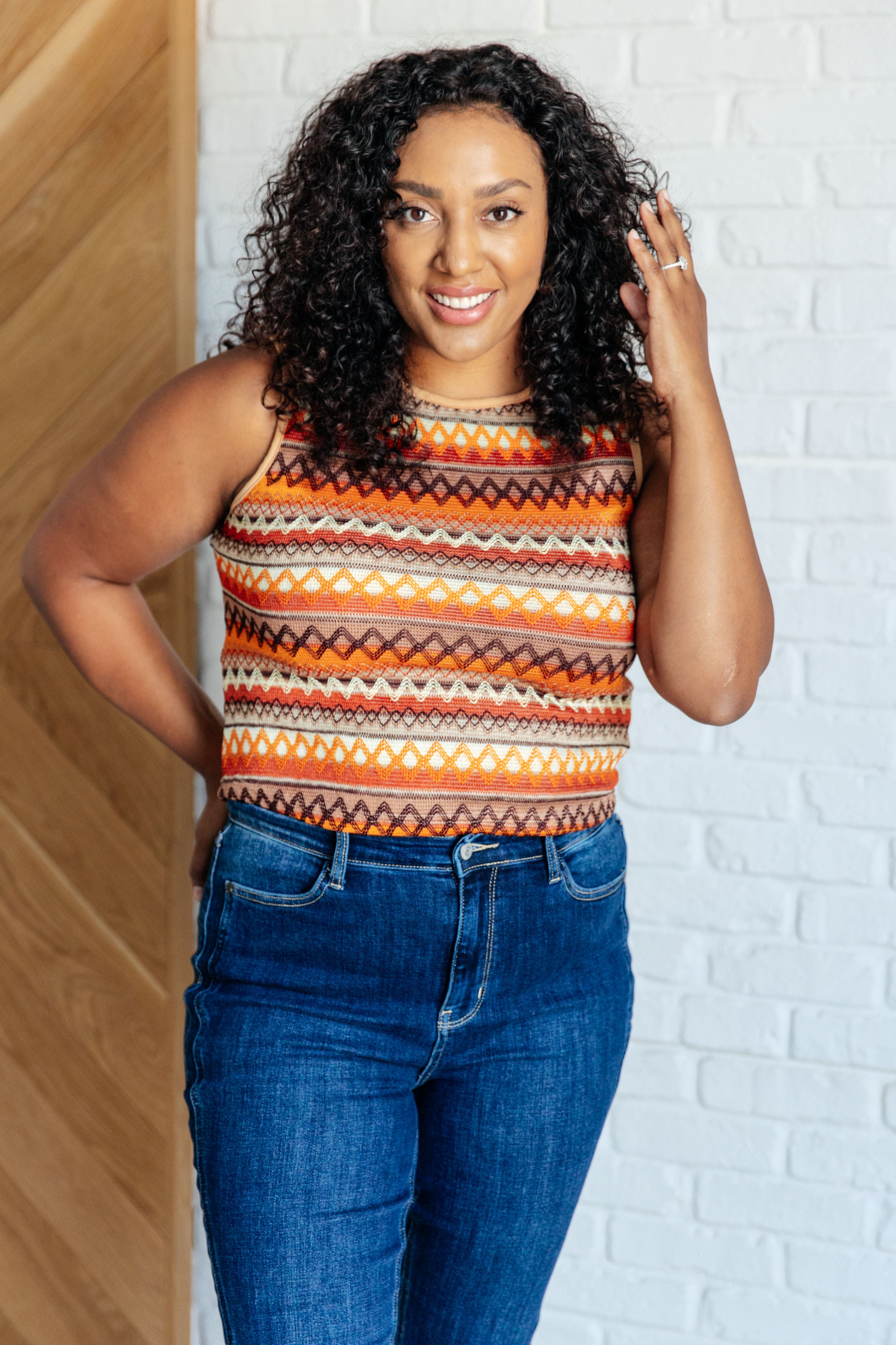 Woman wearing an orange patterned knit tank top paired with high-waisted blue jeans standing indoors smiling