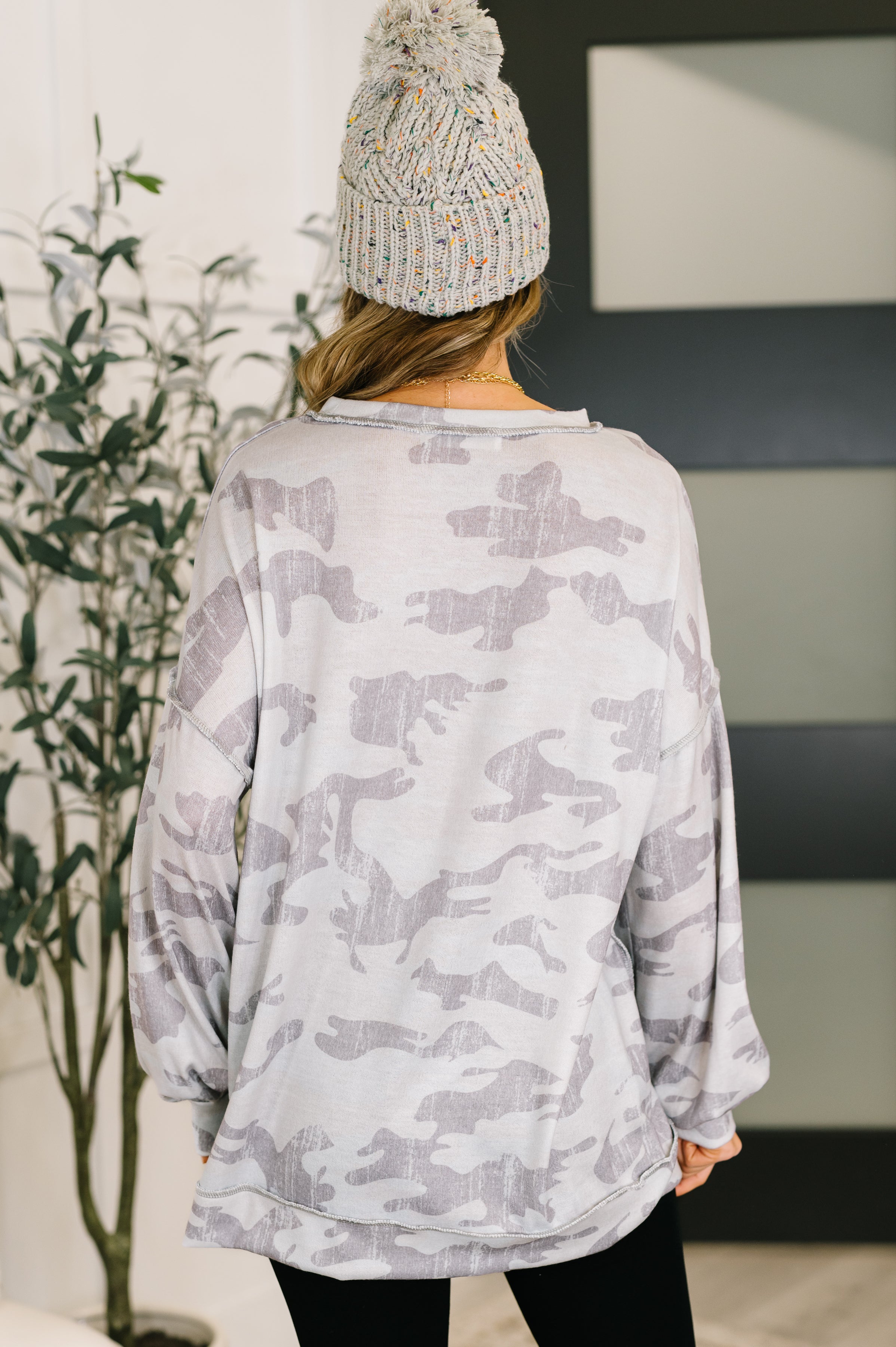 Woman wearing a loose grey camo drop shoulder top with a knit speckled beanie, standing indoors near a plant and door