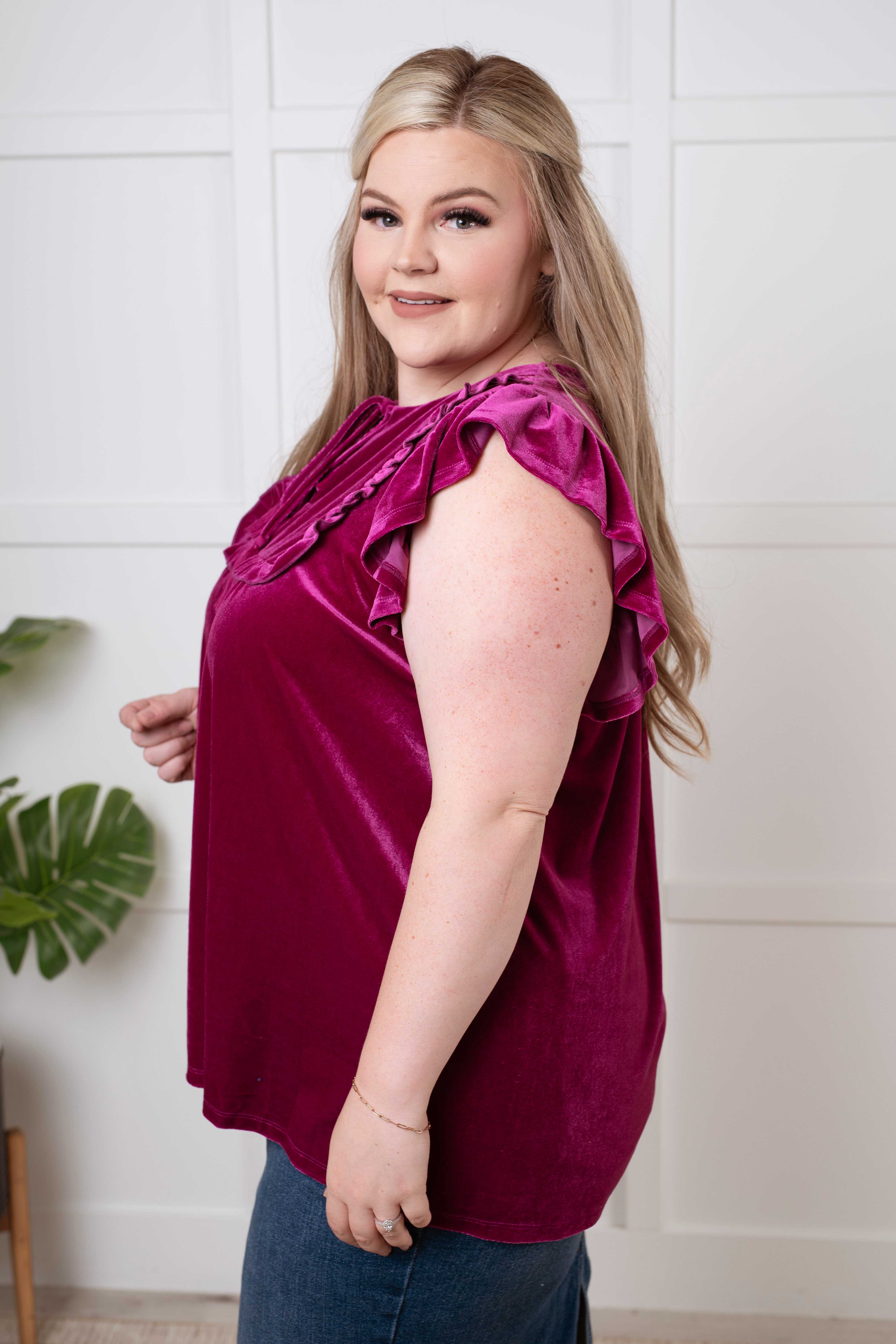 Plus-size model wearing a plush magenta blouse with ruffled cap sleeves standing indoors by a plant