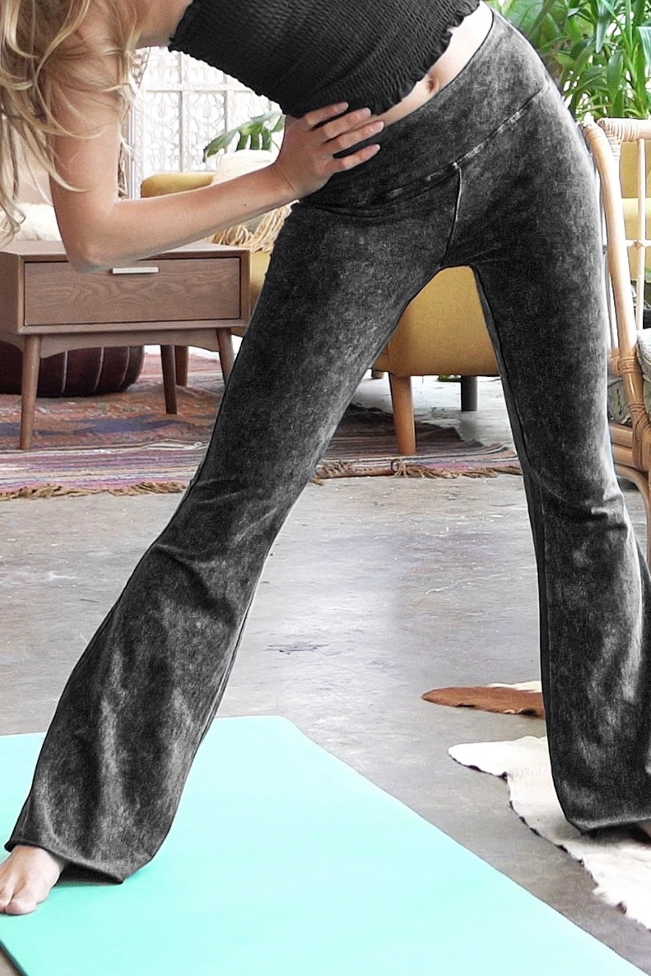 Woman wearing ash black acid wash flare pants stretching on a yoga mat in a bright, bohemian indoor space
