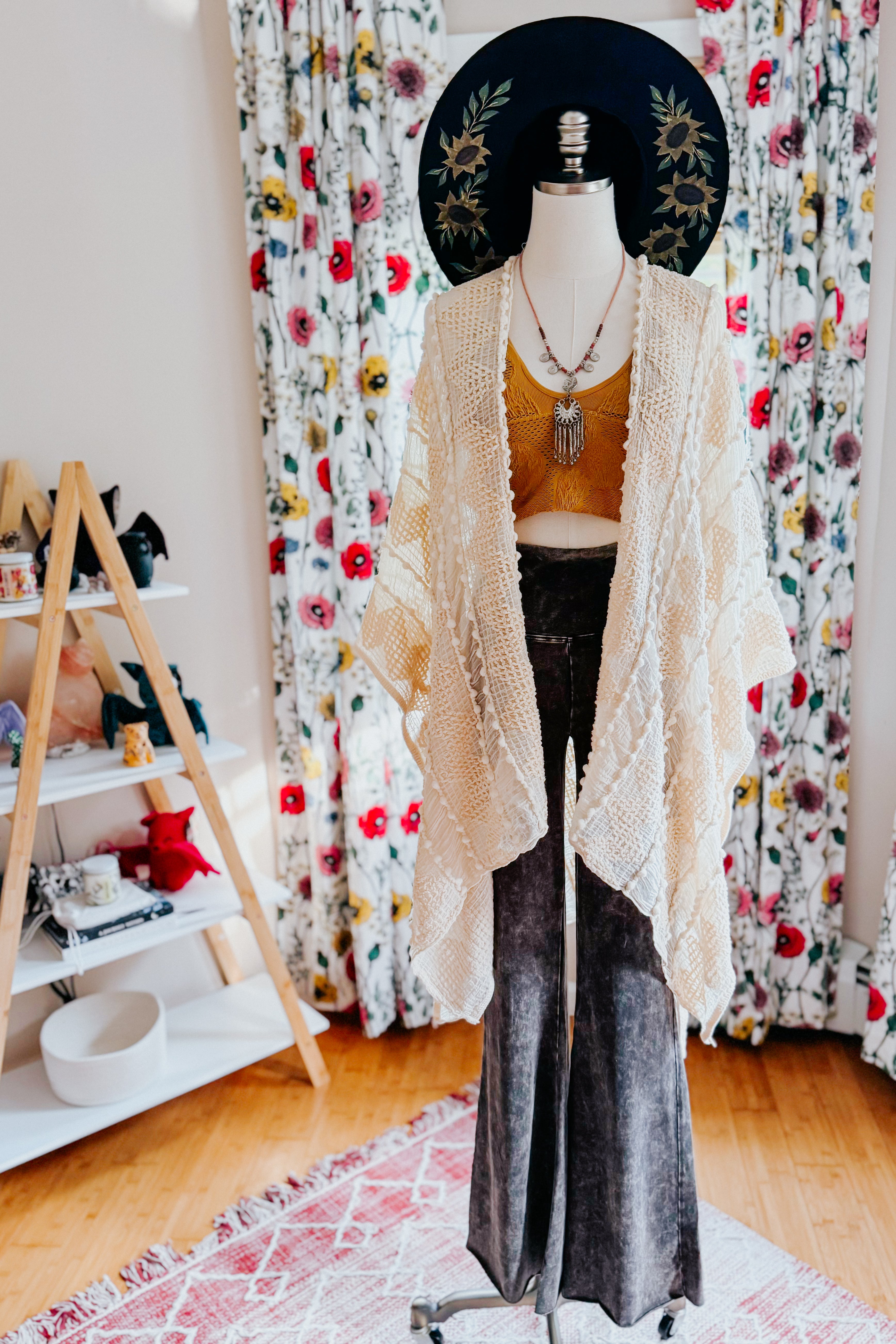 Mannequin dressed in ash black acid wash flared pants, yellow cropped top, cream crochet shawl, and black floral hat indoors