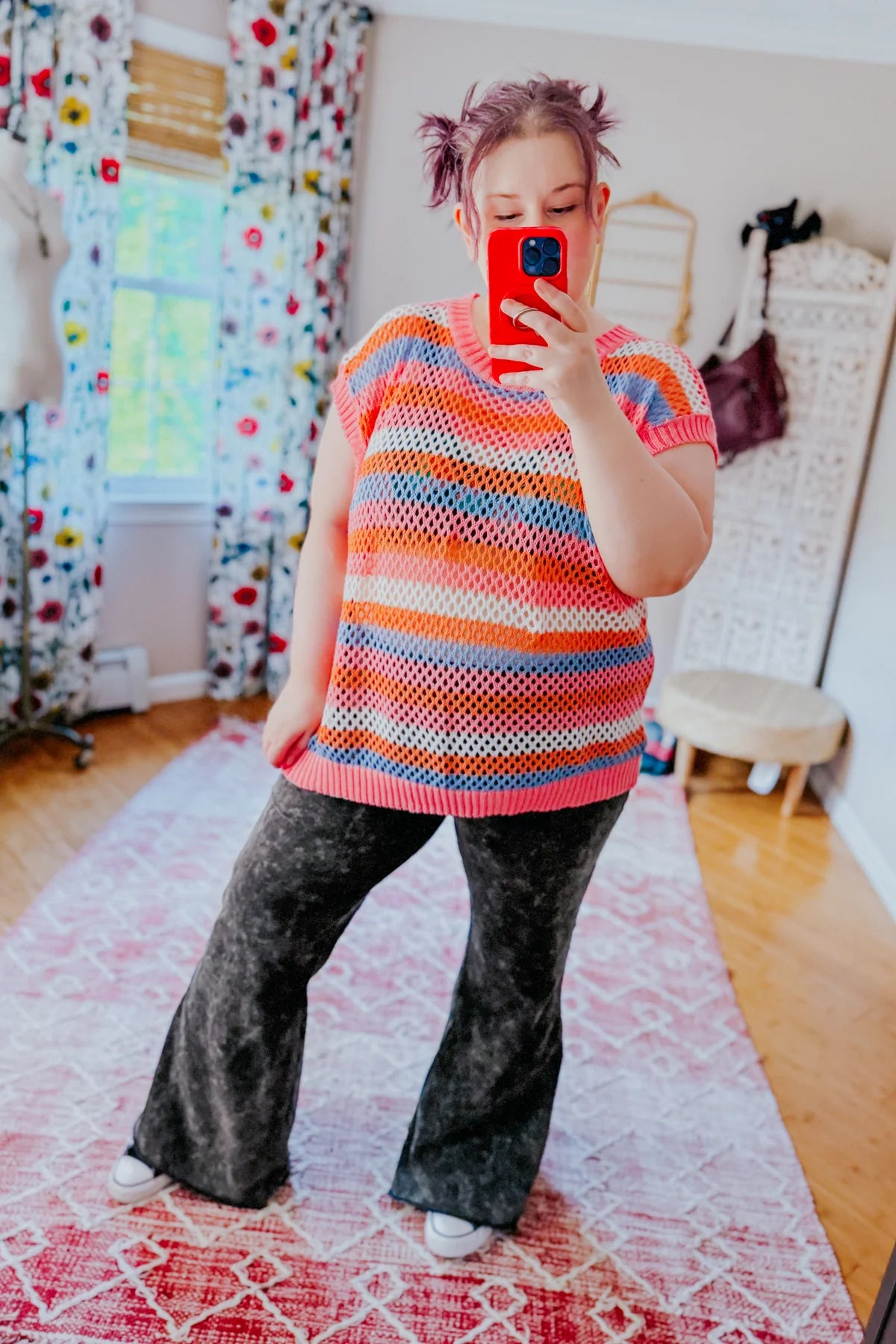 Woman wearing colorful striped knit top with black acid wash flared pants taking mirror selfie indoors