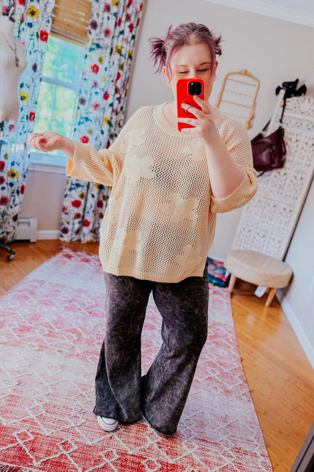 Woman taking mirror selfie wearing cream open-knit sweater with floral patterns and acid wash flared pants indoors
