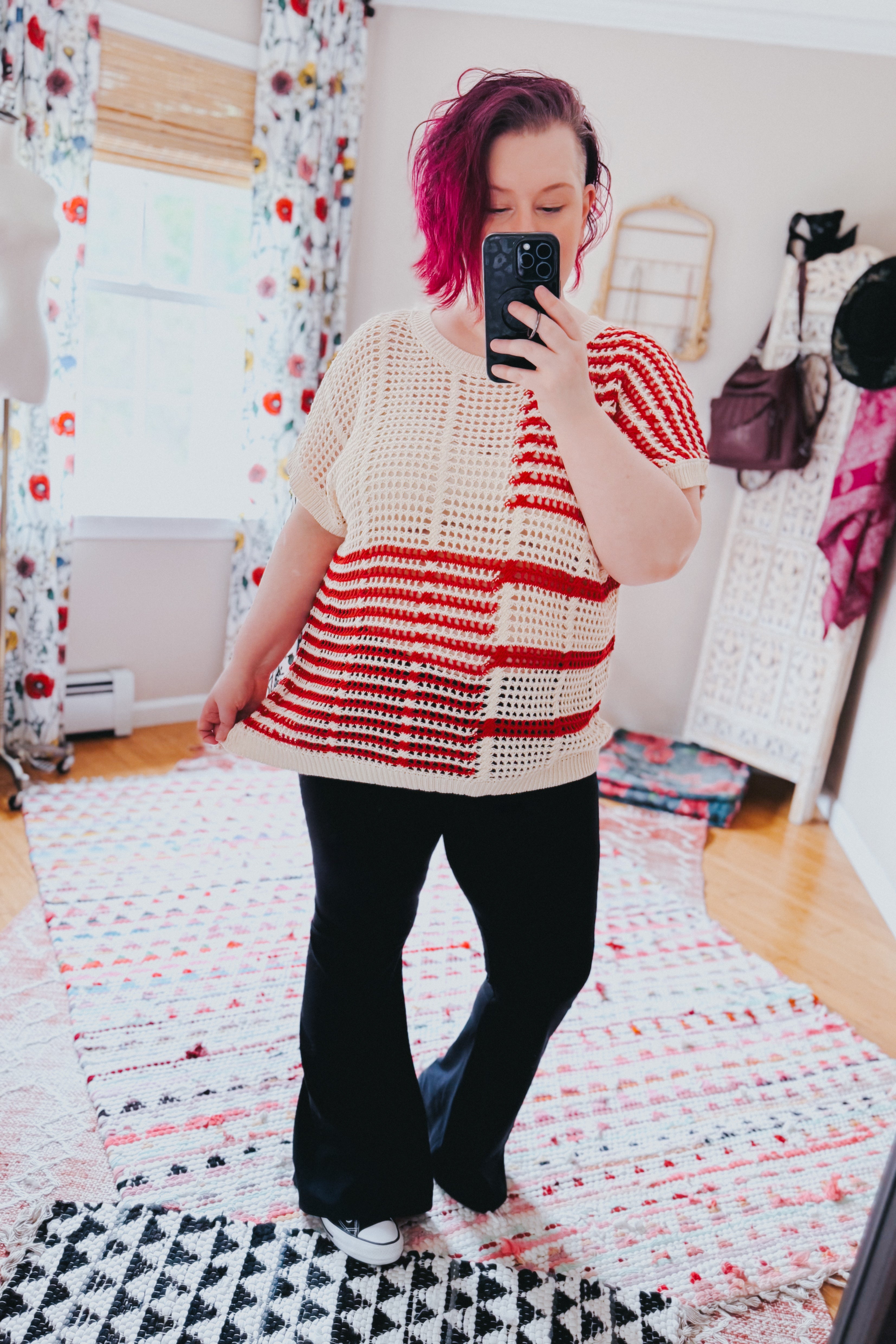 Woman with magenta hair wearing red striped net tee and black flare pants taking mirror selfie in cozy, eclectic room