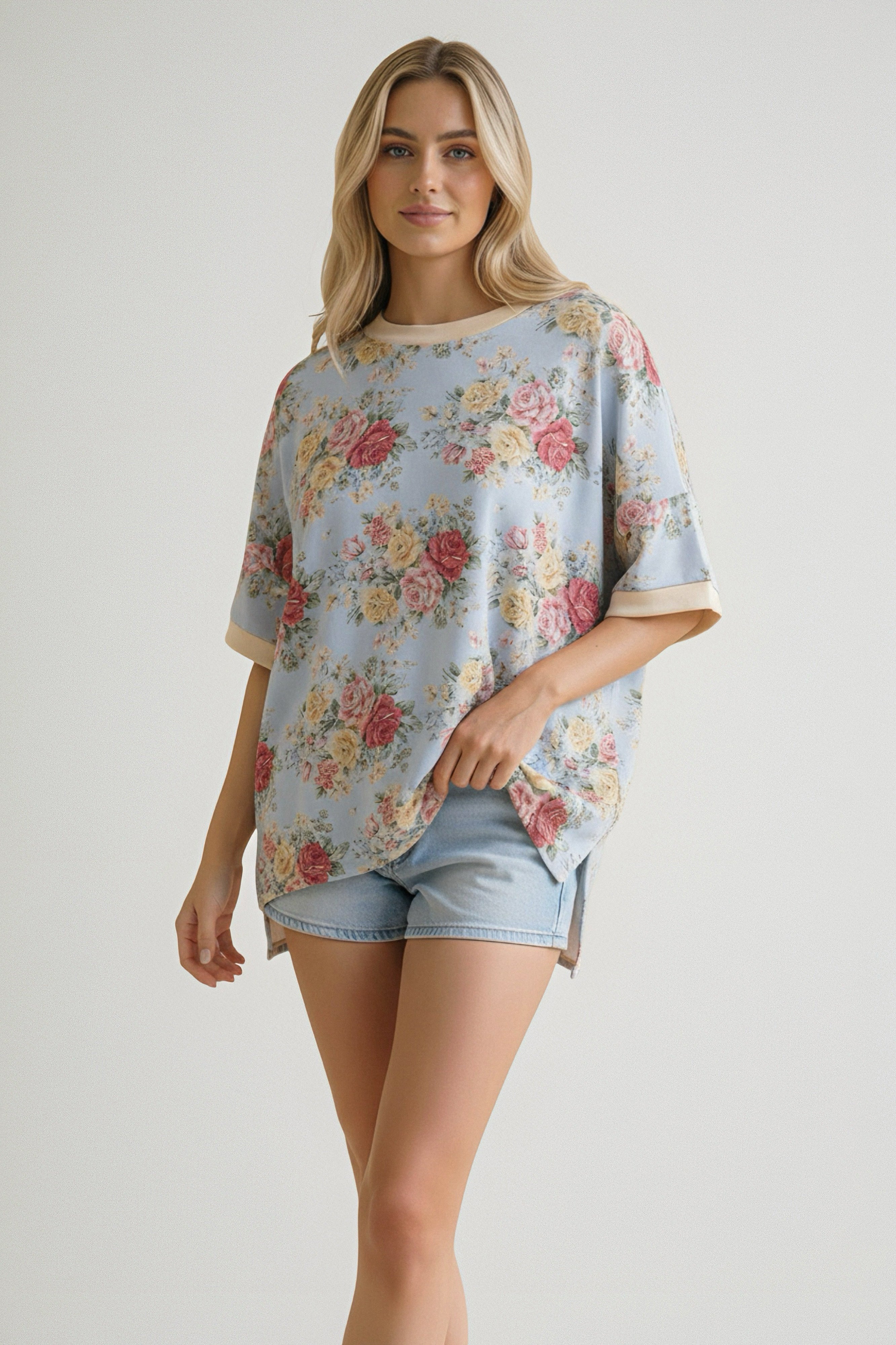 Woman wearing a loose blue top with pink and yellow floral print paired with light denim shorts posing against a plain background