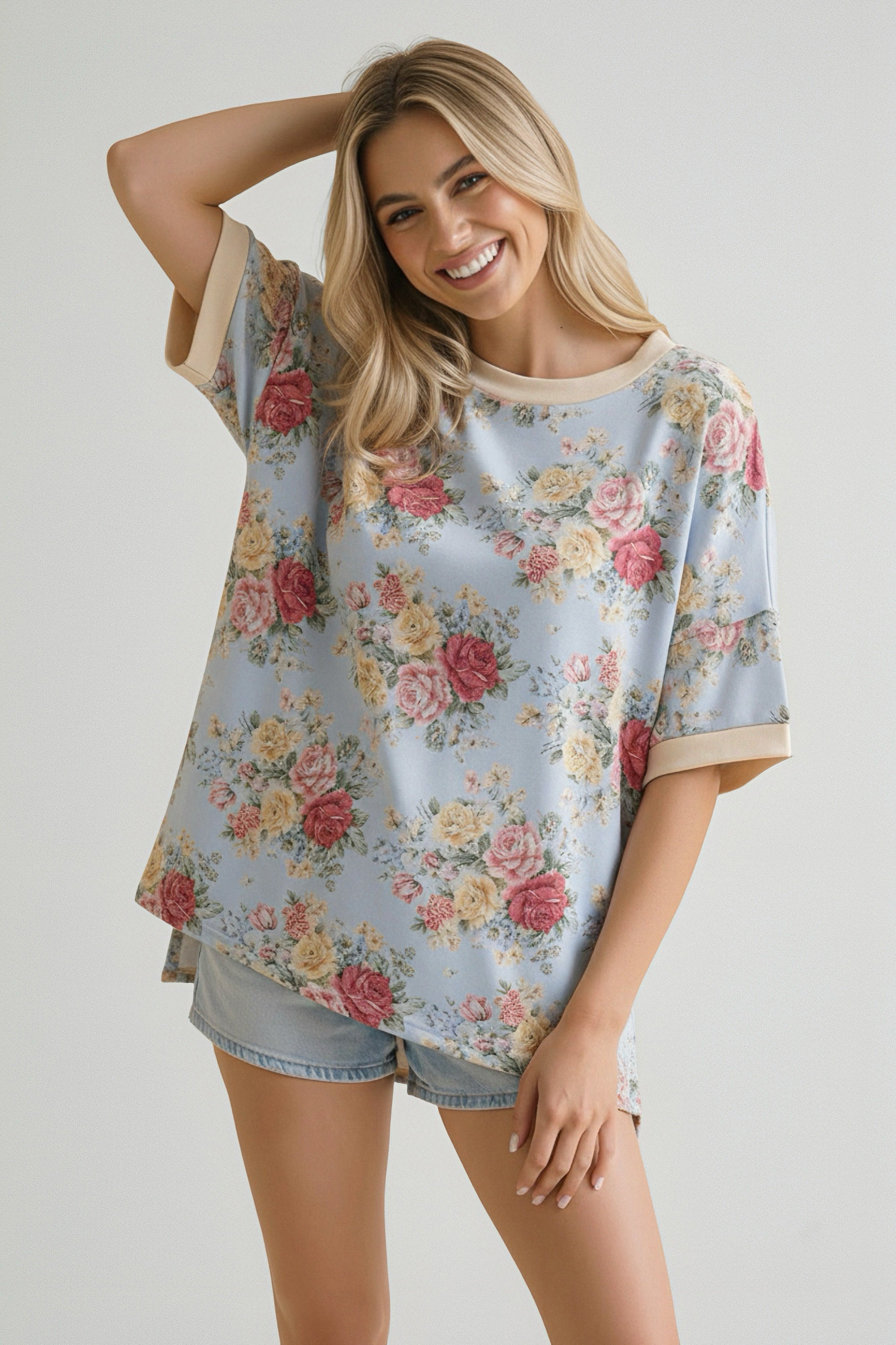 Smiling woman wearing a loose blue floral top with beige trim and light denim shorts, posing with hand behind head