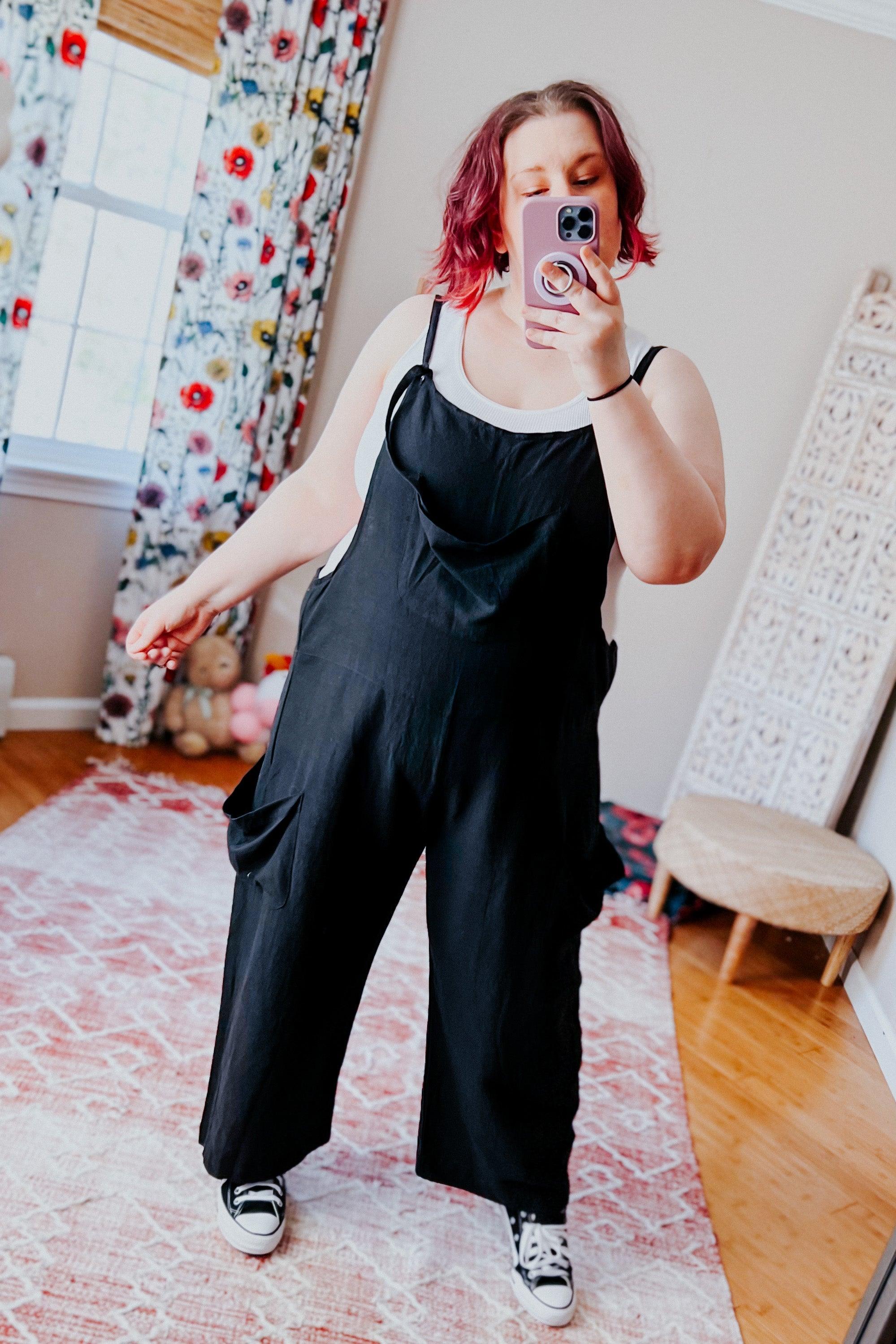 Plus-size woman taking mirror selfie wearing black slouchy overalls layered over white tank top with sneakers indoors