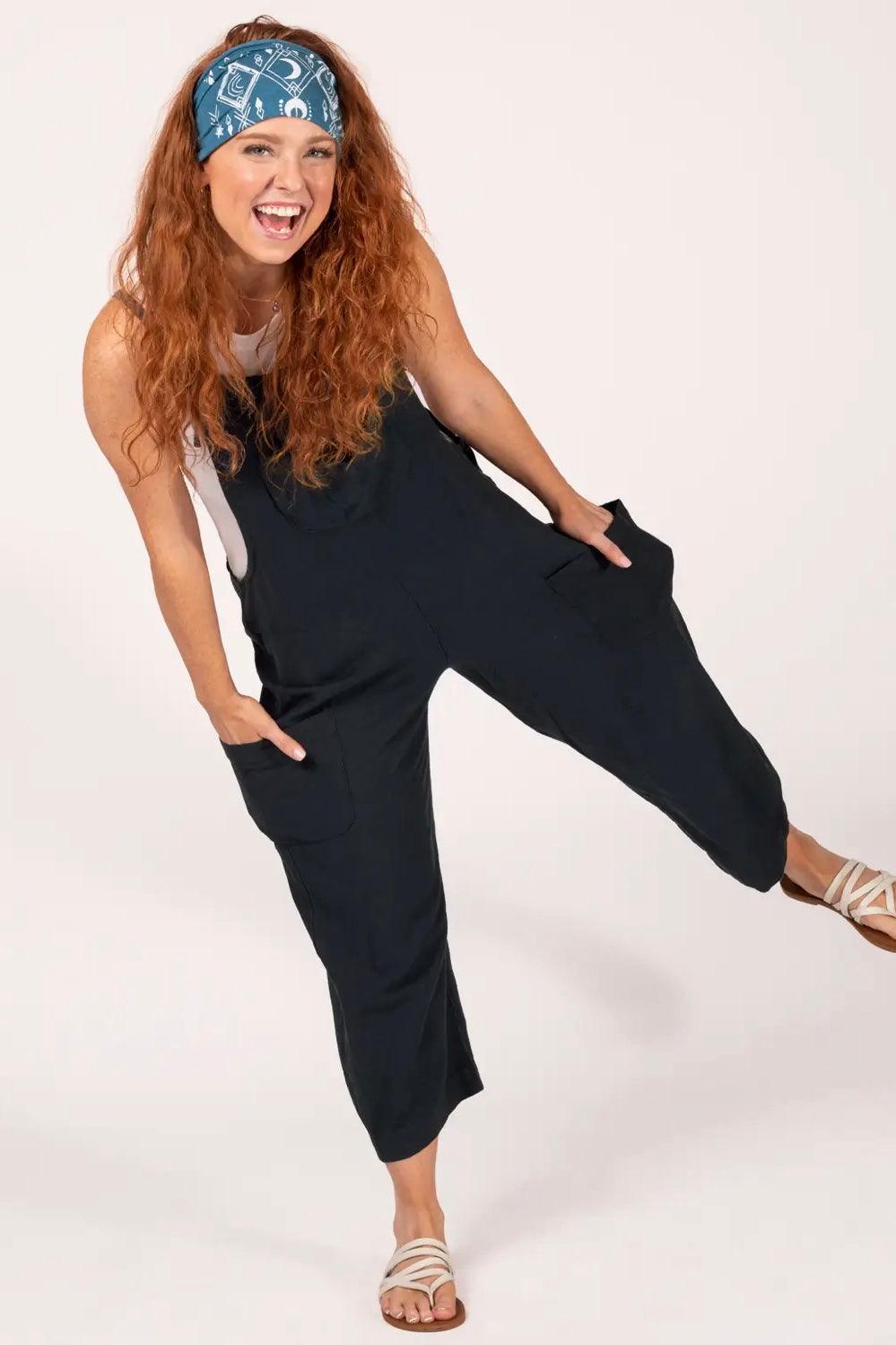 Woman joyfully posing in black slouchy overalls with large pockets, white tank top, blue patterned headband, and sandals