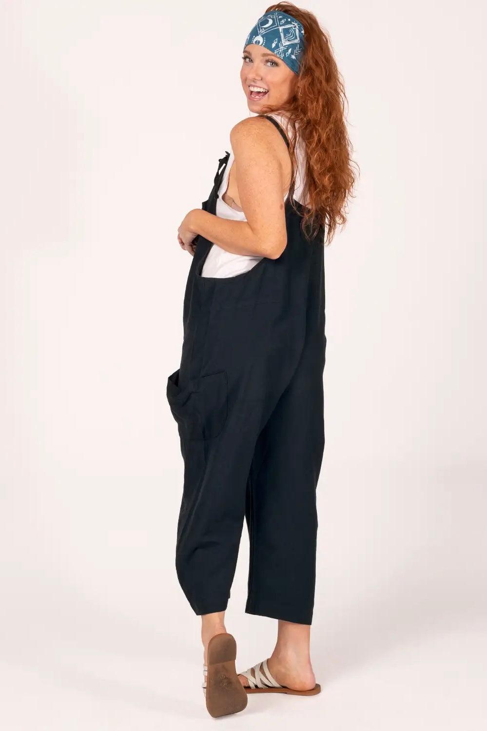 Smiling woman in black slouchy overalls with side pockets, white tank top, sandals, and blue patterned headband