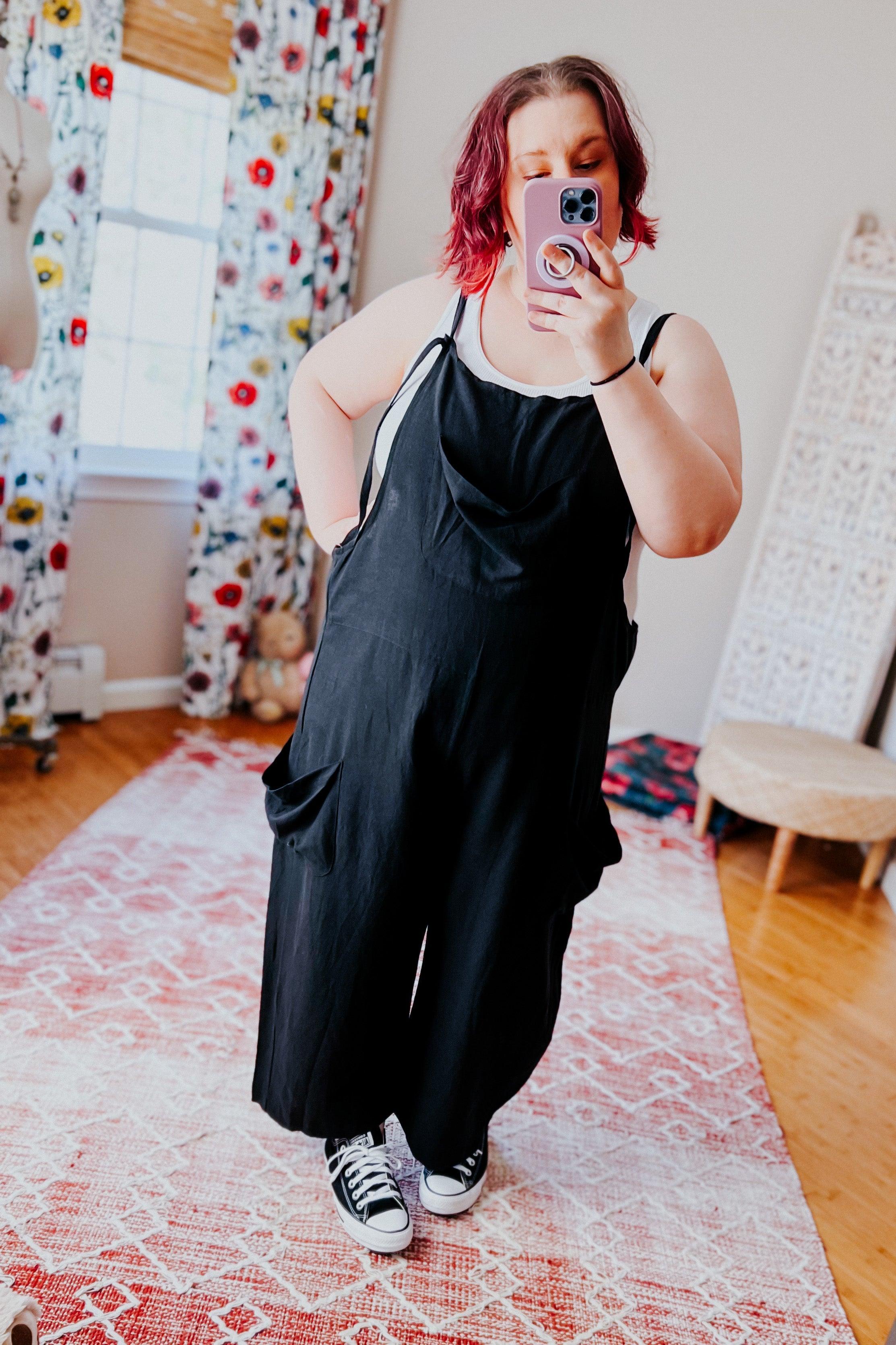 Woman wearing loose black overalls with white tank top and black sneakers taking selfie indoors on patterned rug