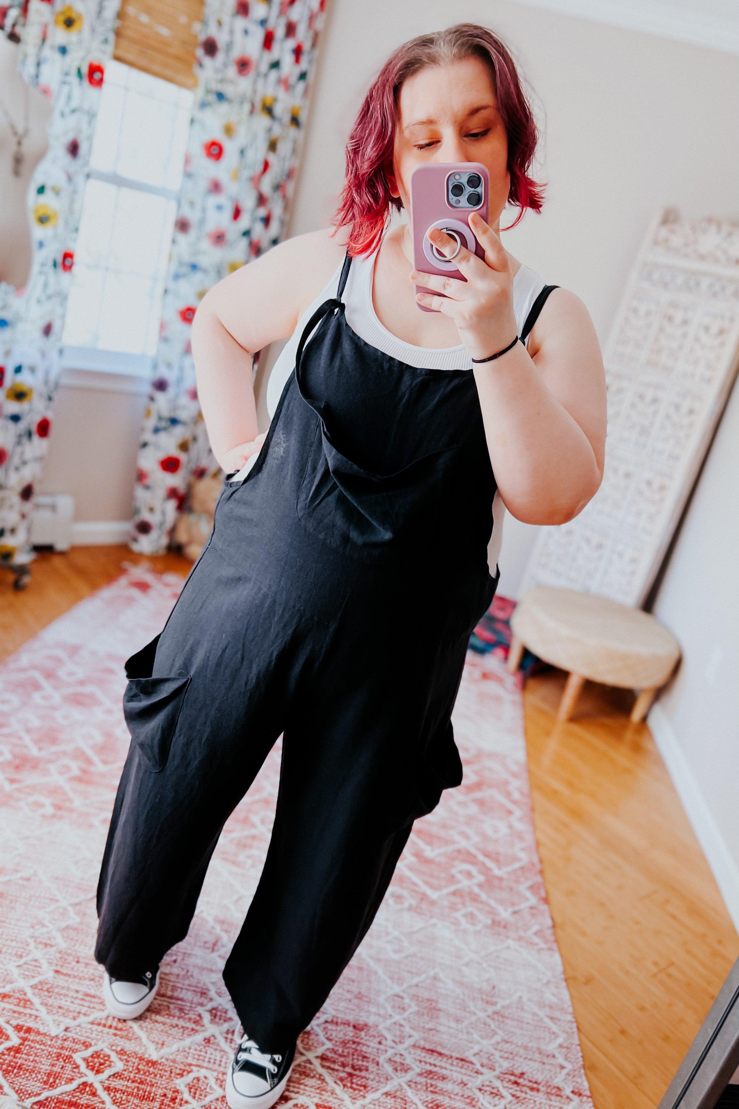 Woman wearing black slouchy overalls with white tank top and black sneakers taking a mirror selfie indoors