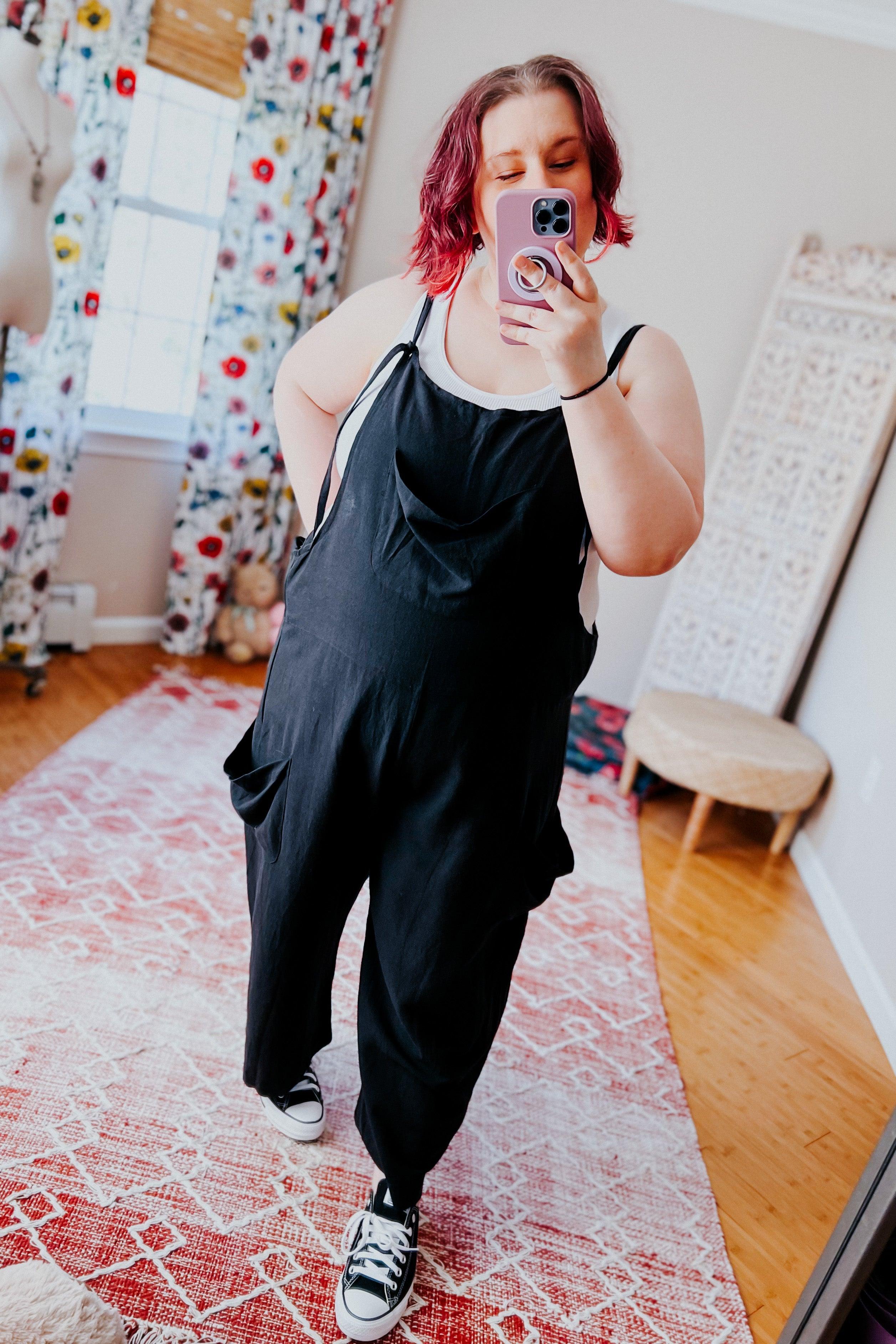 Plus size woman wearing black slouchy overalls with white tank top and sneakers taking mirror selfie indoors