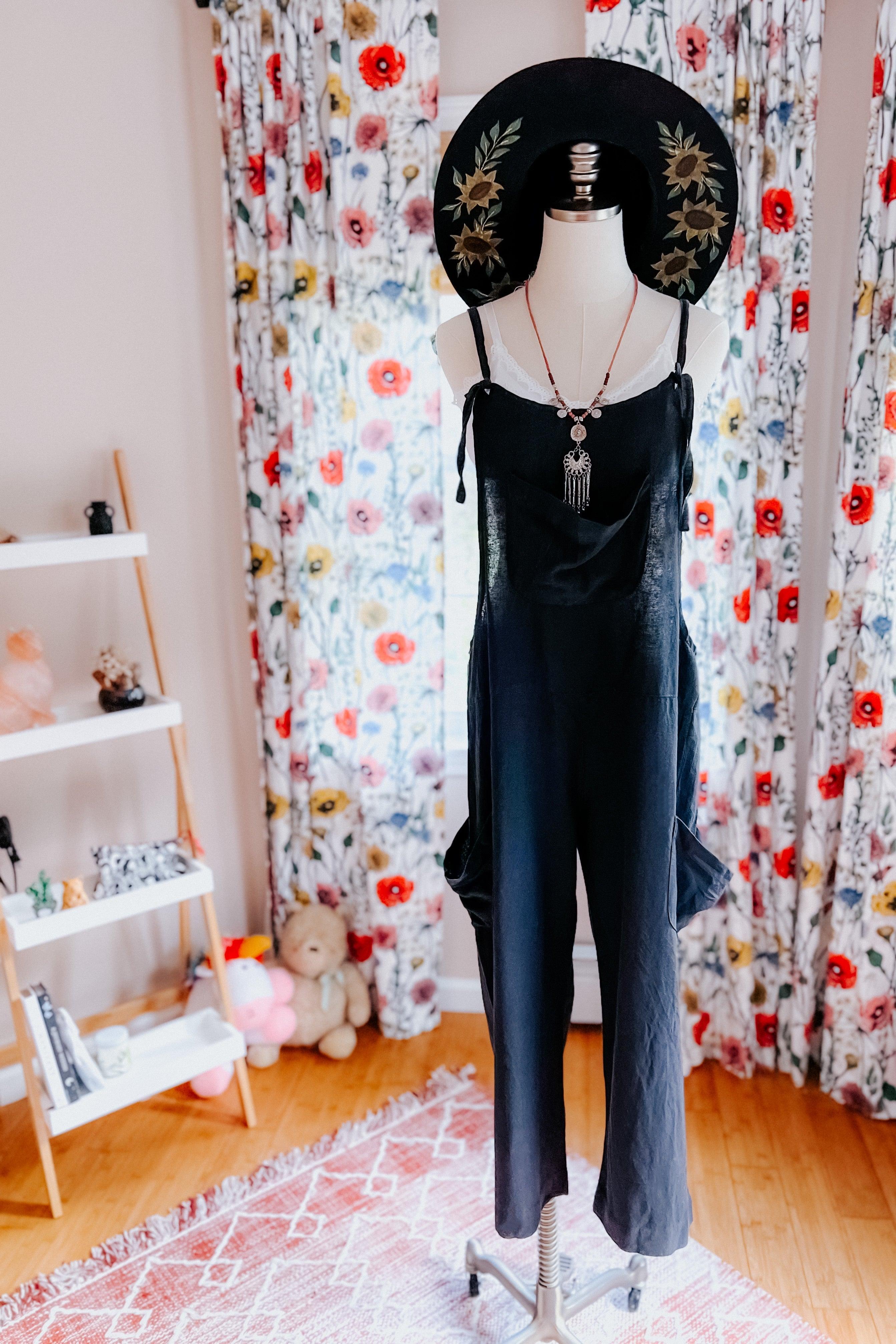 Mannequin dressed in black slouchy overalls with layered necklaces and wide-brimmed hat in cozy room setting