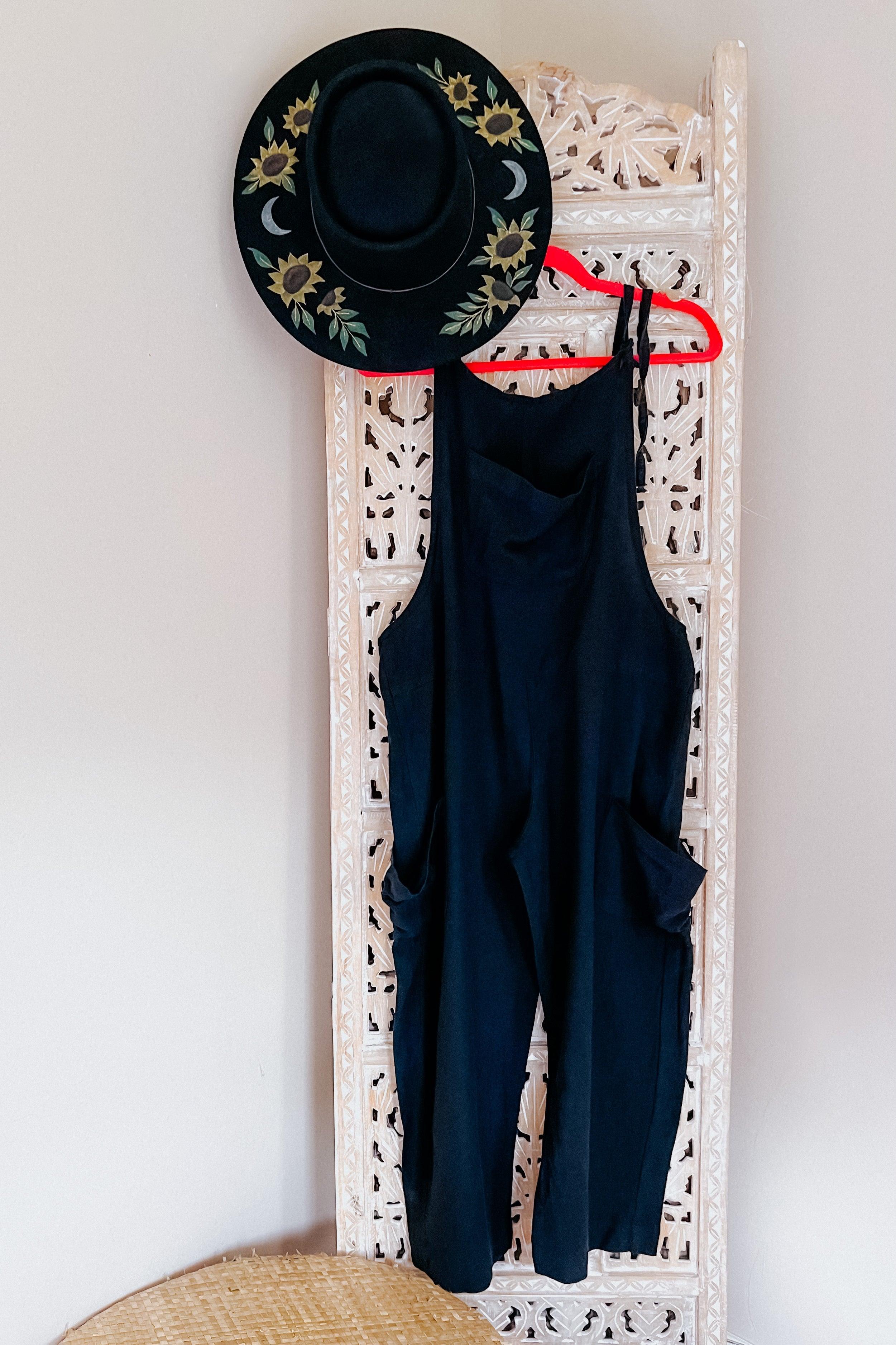 Black slouchy overalls hanging on a carved wooden screen next to a black hat with sunflower and moon designs