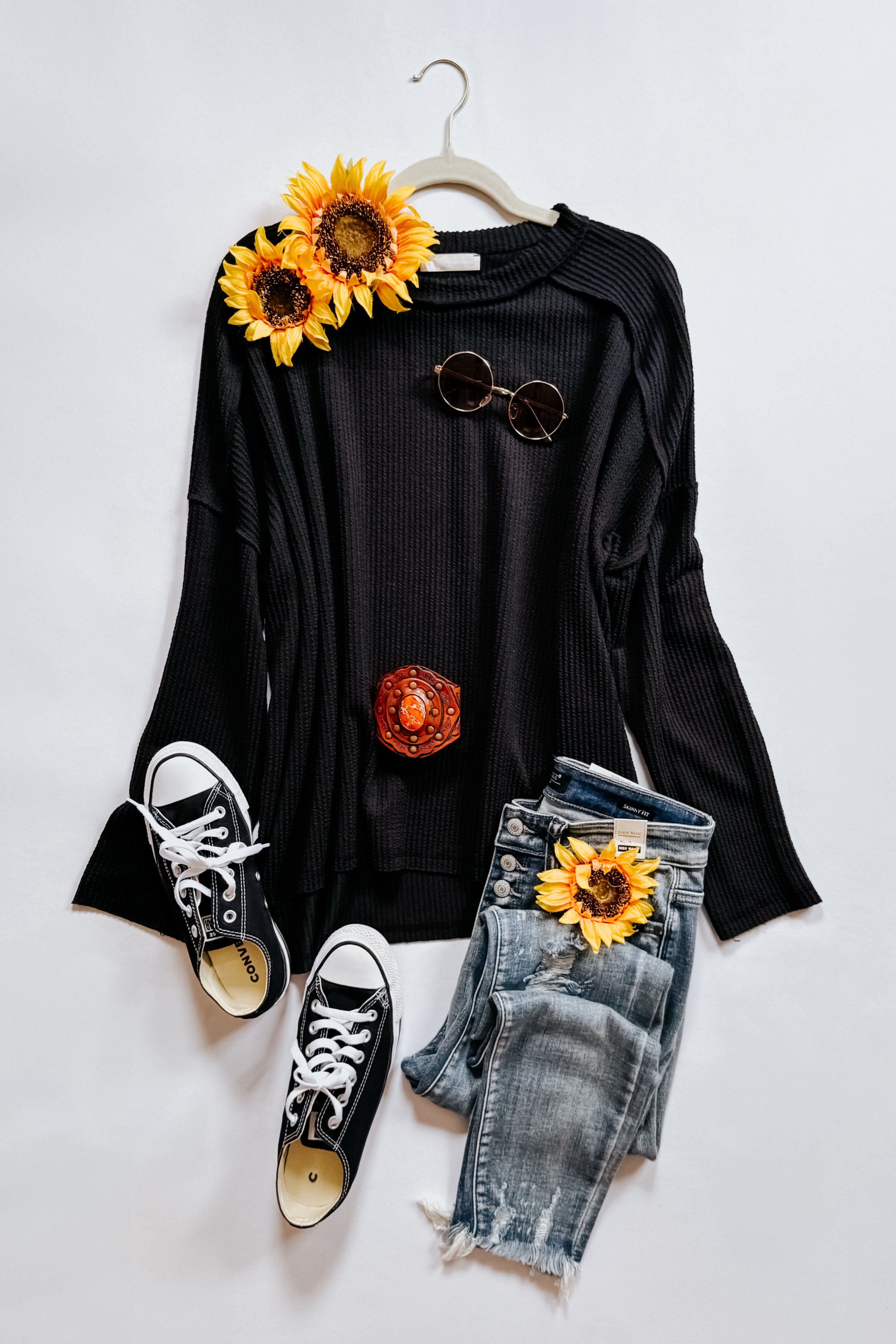 Black ribbed knit drop shoulder top styled with distressed jeans, black sneakers, round sunglasses, and sunflowers on white background