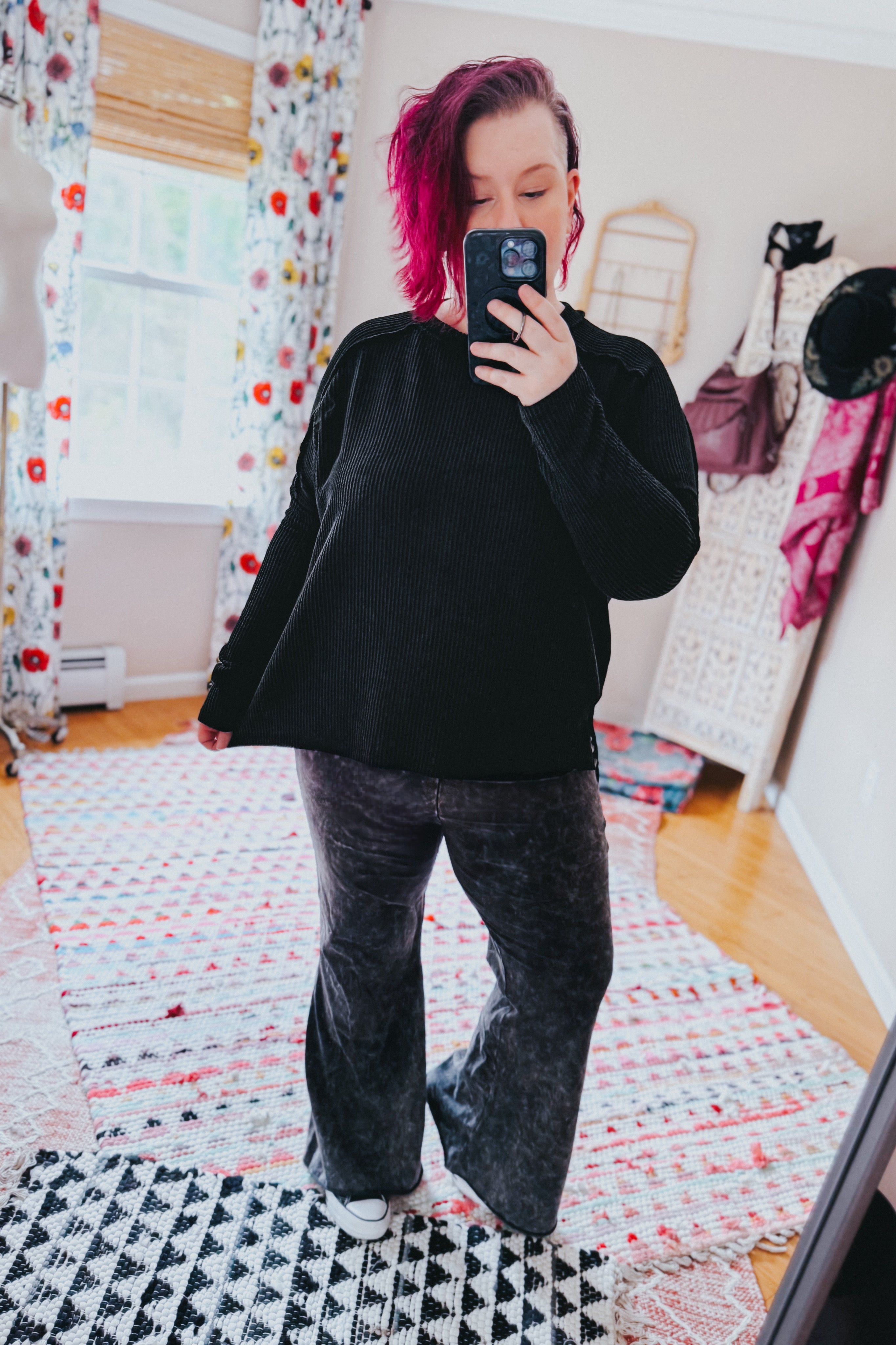 Woman with short pink hair wearing black cable ribbed knit top and dark velvet pants taking a mirror selfie indoors