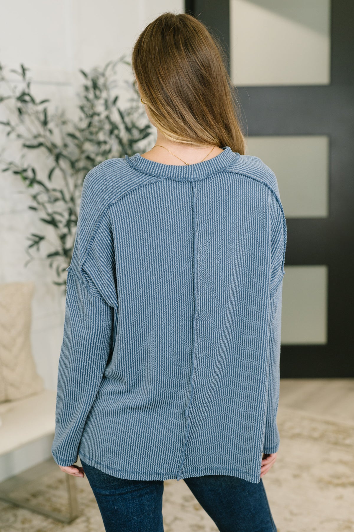 Woman viewed from behind wearing a dusty blue cable ribbed drop shoulder top with jeans in a modern home interior