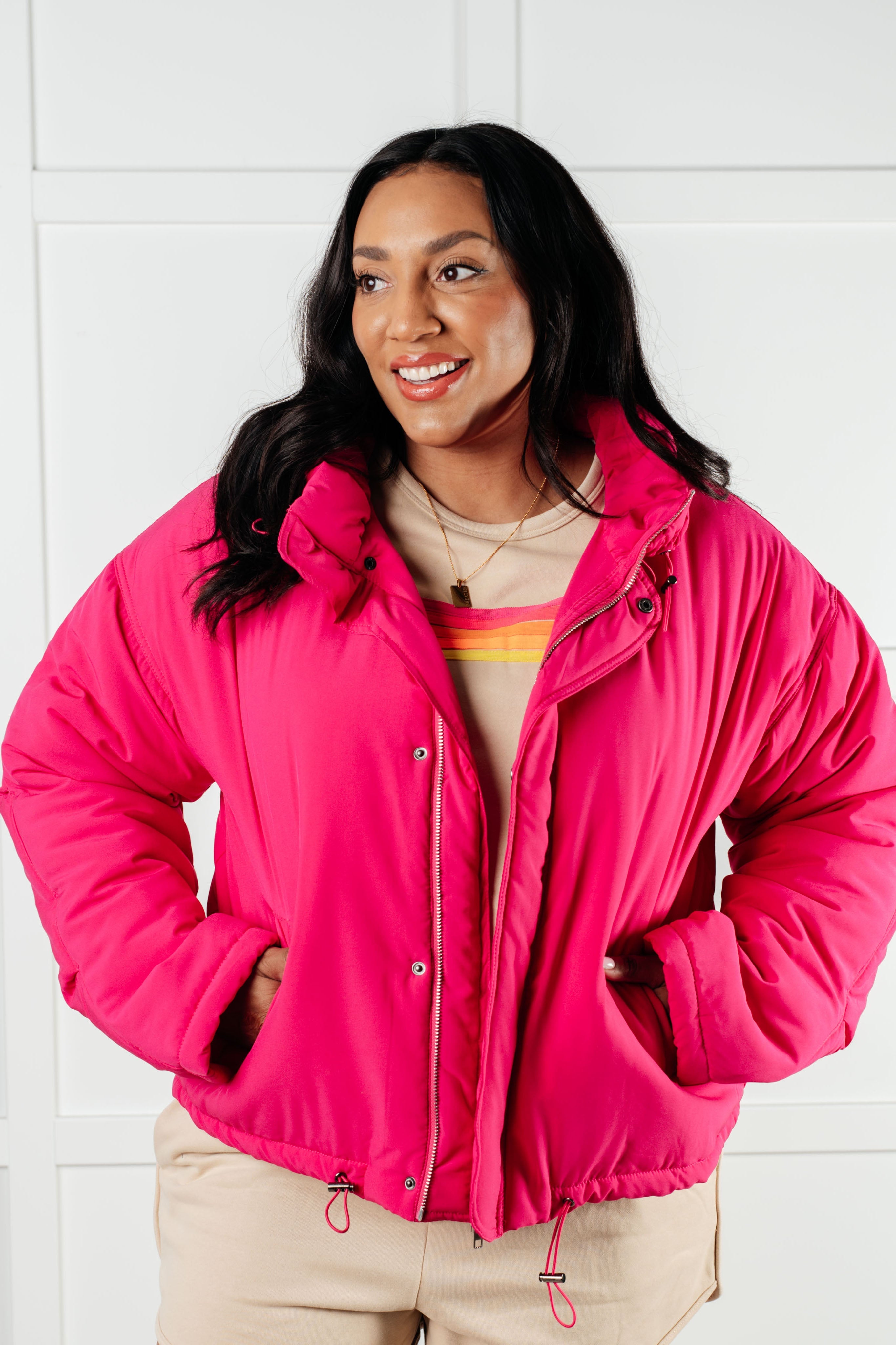 Woman smiling wearing bright pink puffer jacket over beige outfit with colorful striped detail indoors