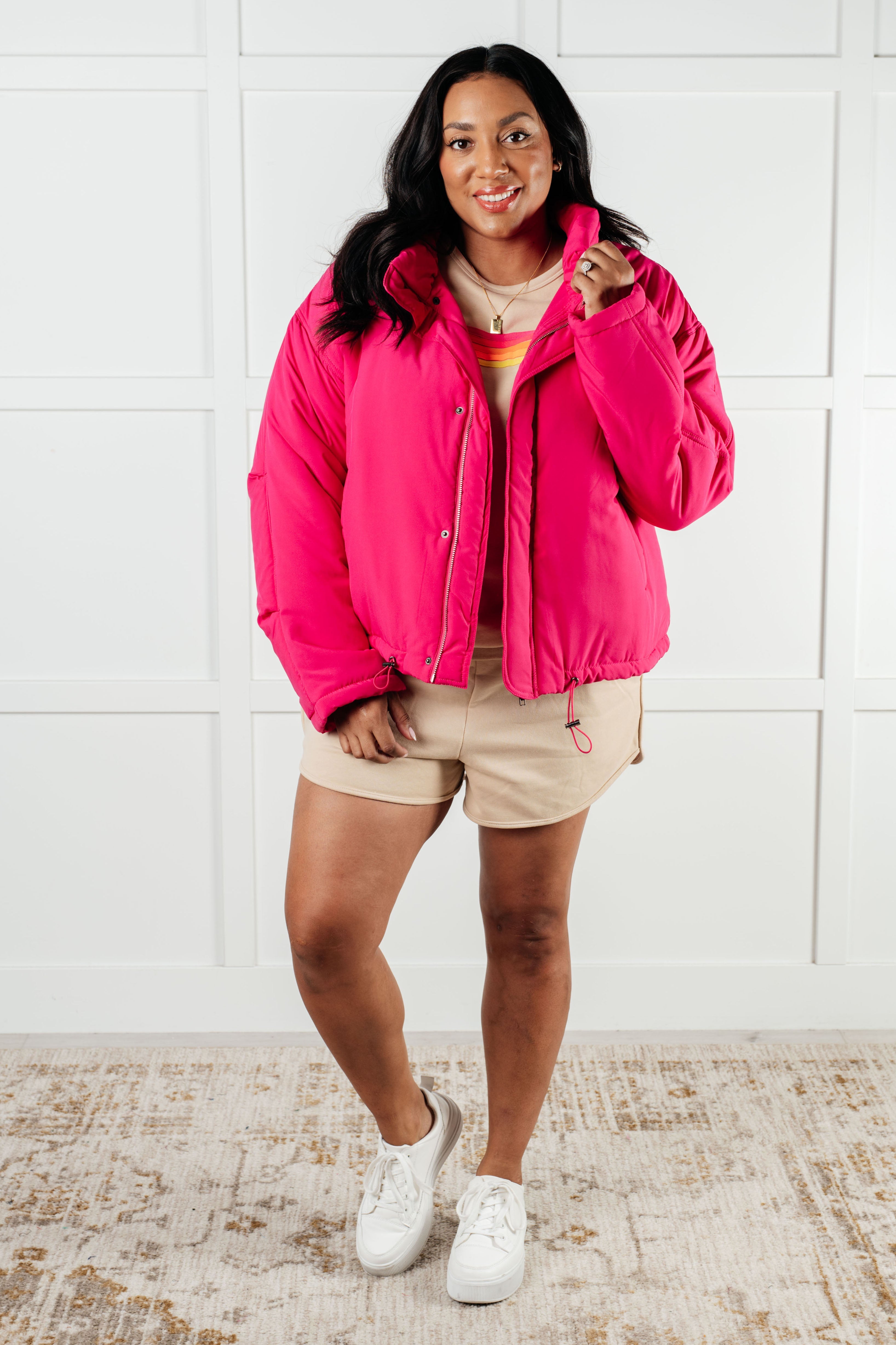Woman wearing bright pink puffer jacket styled with beige shorts and white sneakers indoors on patterned rug