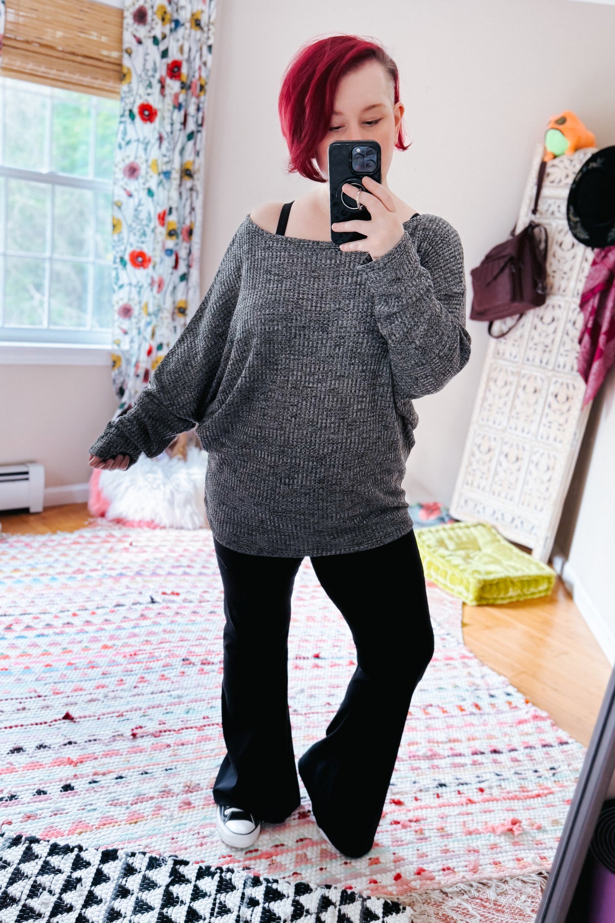 Red haired woman taking a mirror selfie in a charcoal waffle knit boat neck top with dolman sleeves black flared pants and sneakers in a bedroom