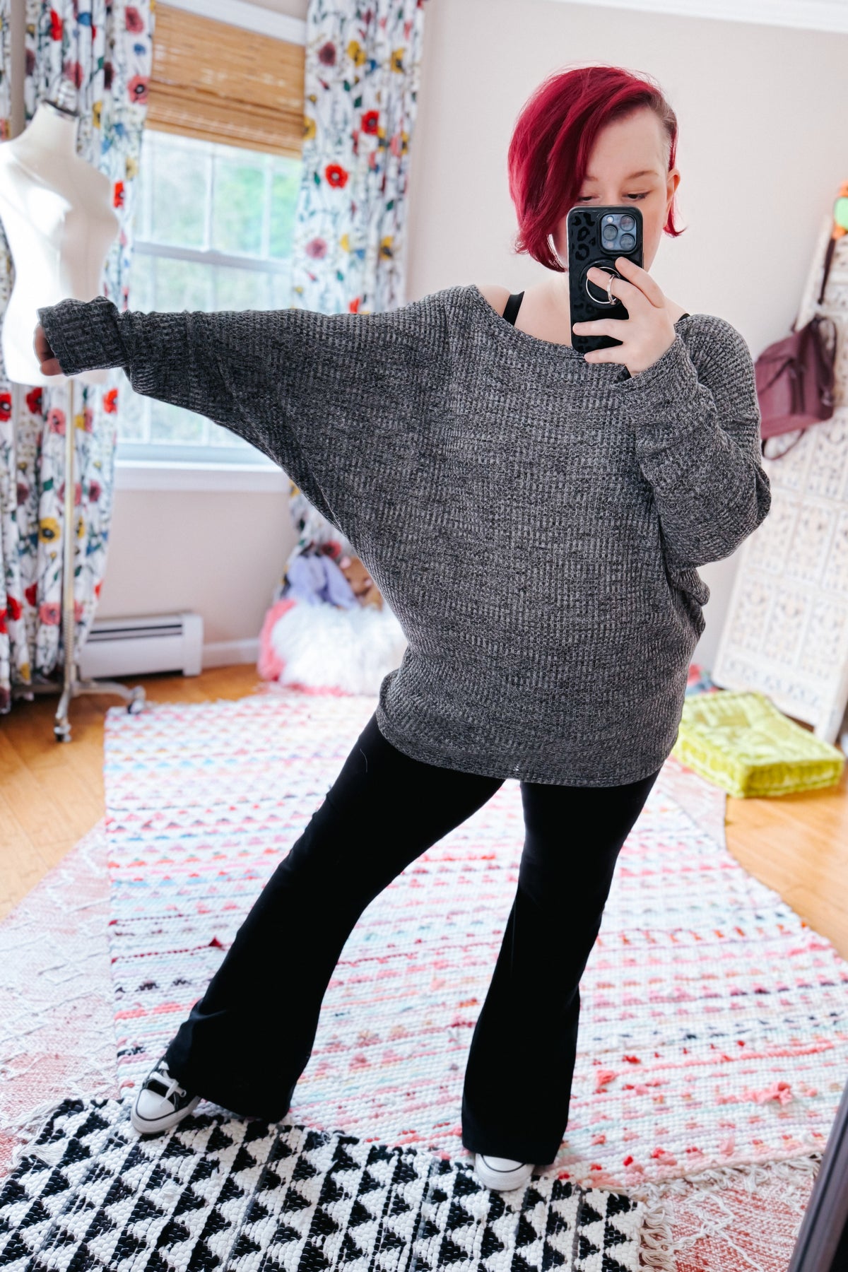 Red haired woman taking a bedroom mirror selfie in a charcoal ribbed top with dolman sleeves and black pants on a patterned rug