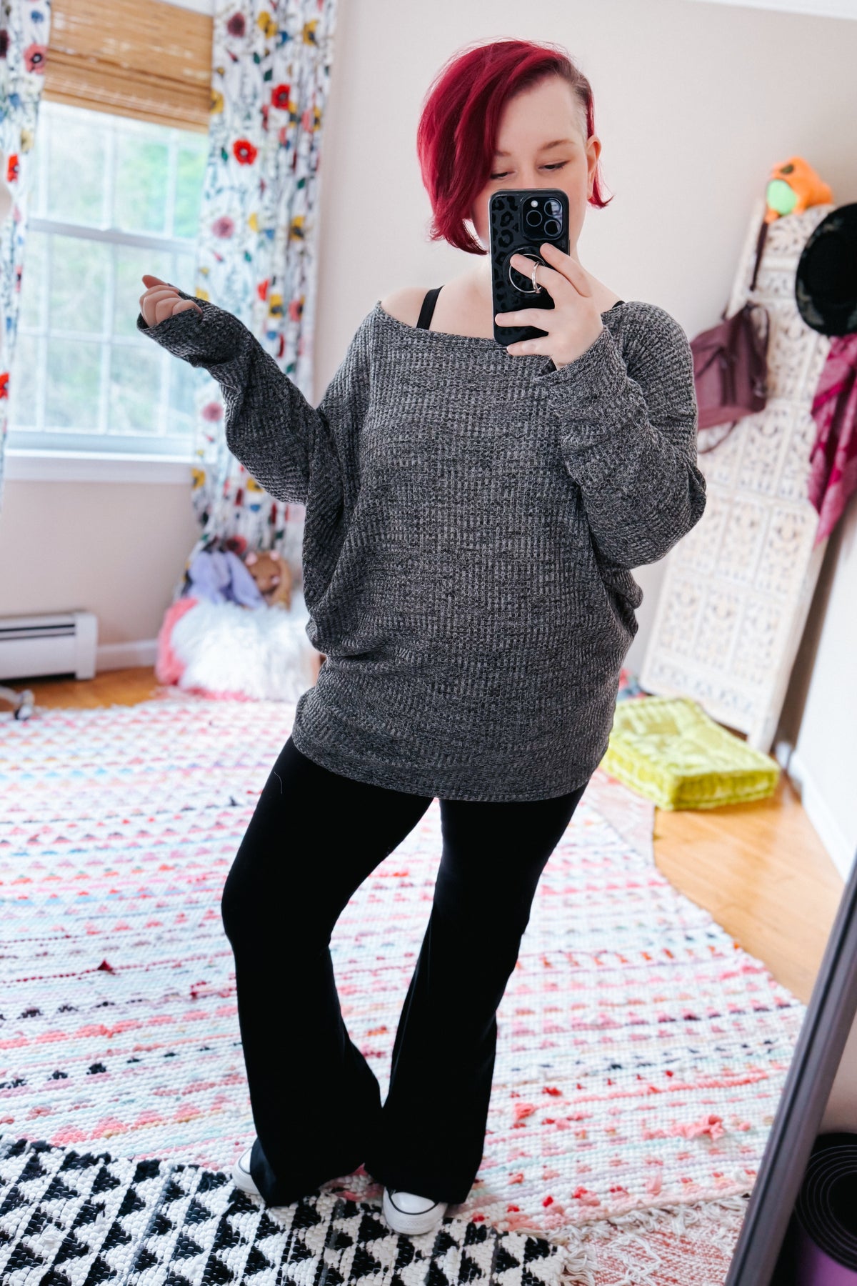 Red haired woman taking a mirror selfie in a charcoal waffle knit boat neck top with dolman sleeves and black pants in a colorful bedroom