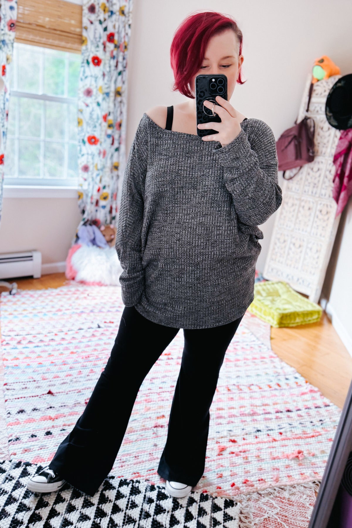 Red haired woman modeling a relaxed charcoal waffle knit boat neck top with dolman sleeves black flared pants and sneakers in a colorful bedroom