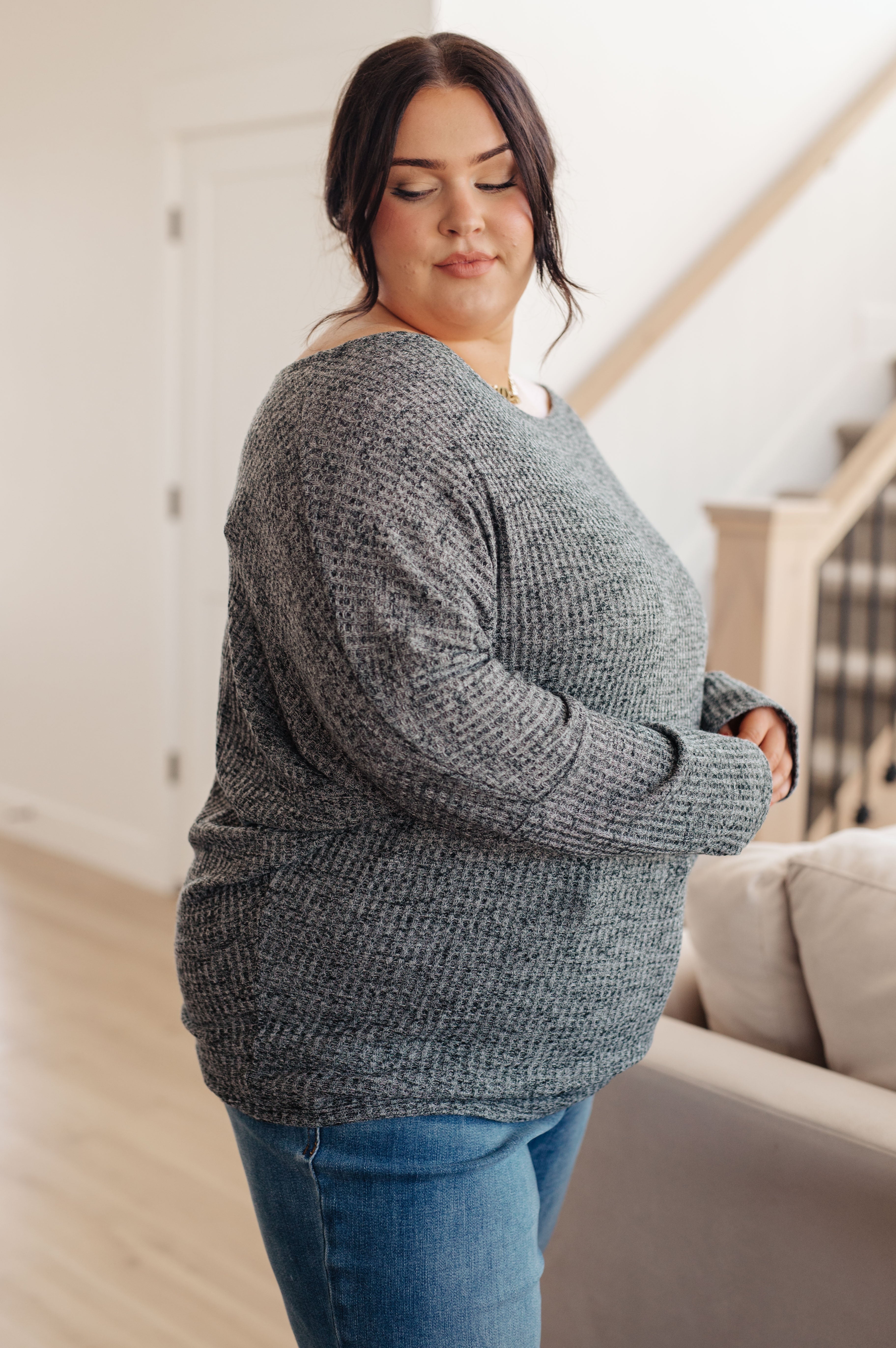 Plus-size woman wearing charcoal ribbed top paired with blue jeans standing indoors near light-colored sofa