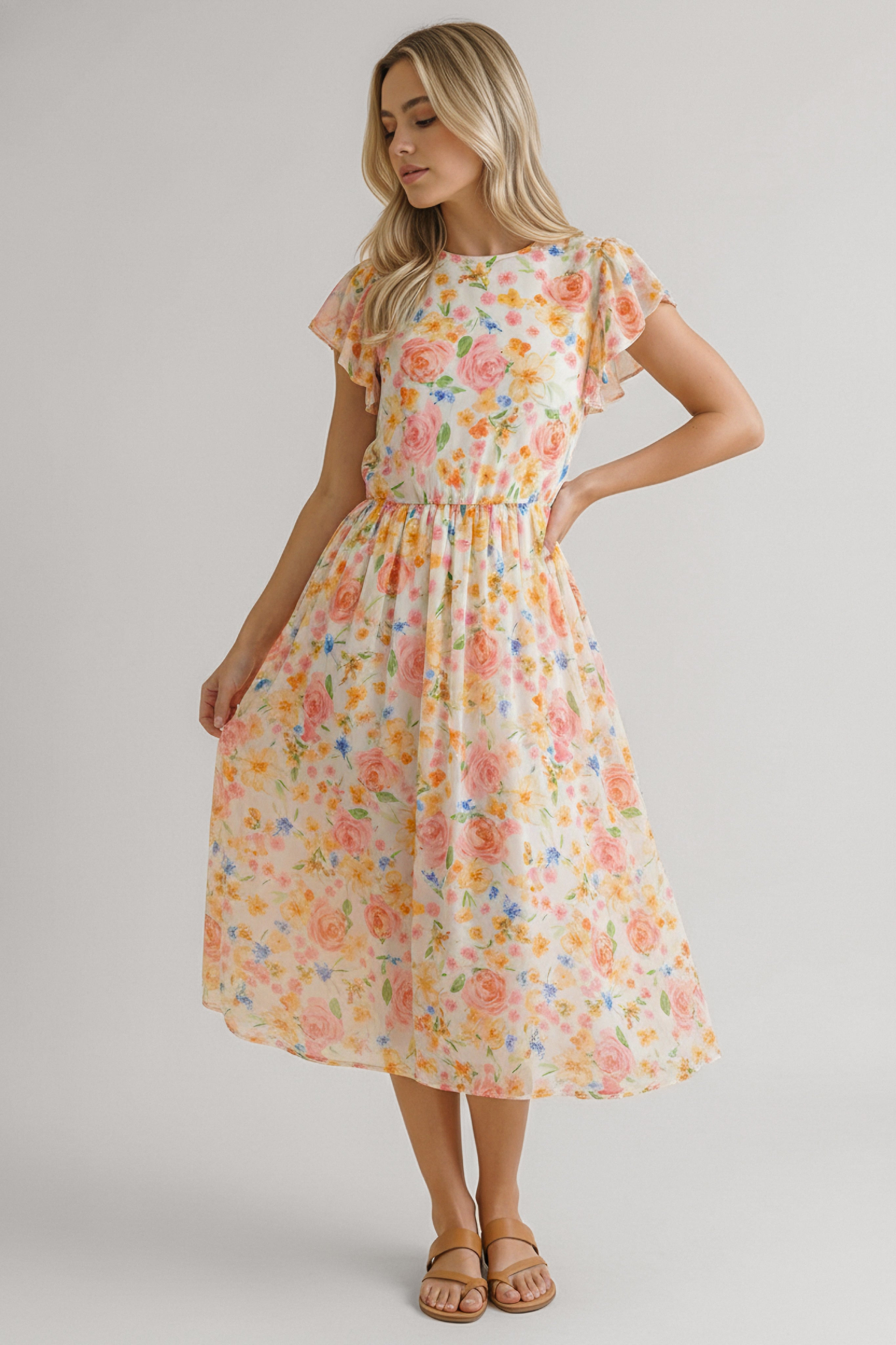 Woman wearing a lightweight midi dress with soft orange and pink floral pattern and flutter sleeves standing gently holding skirt