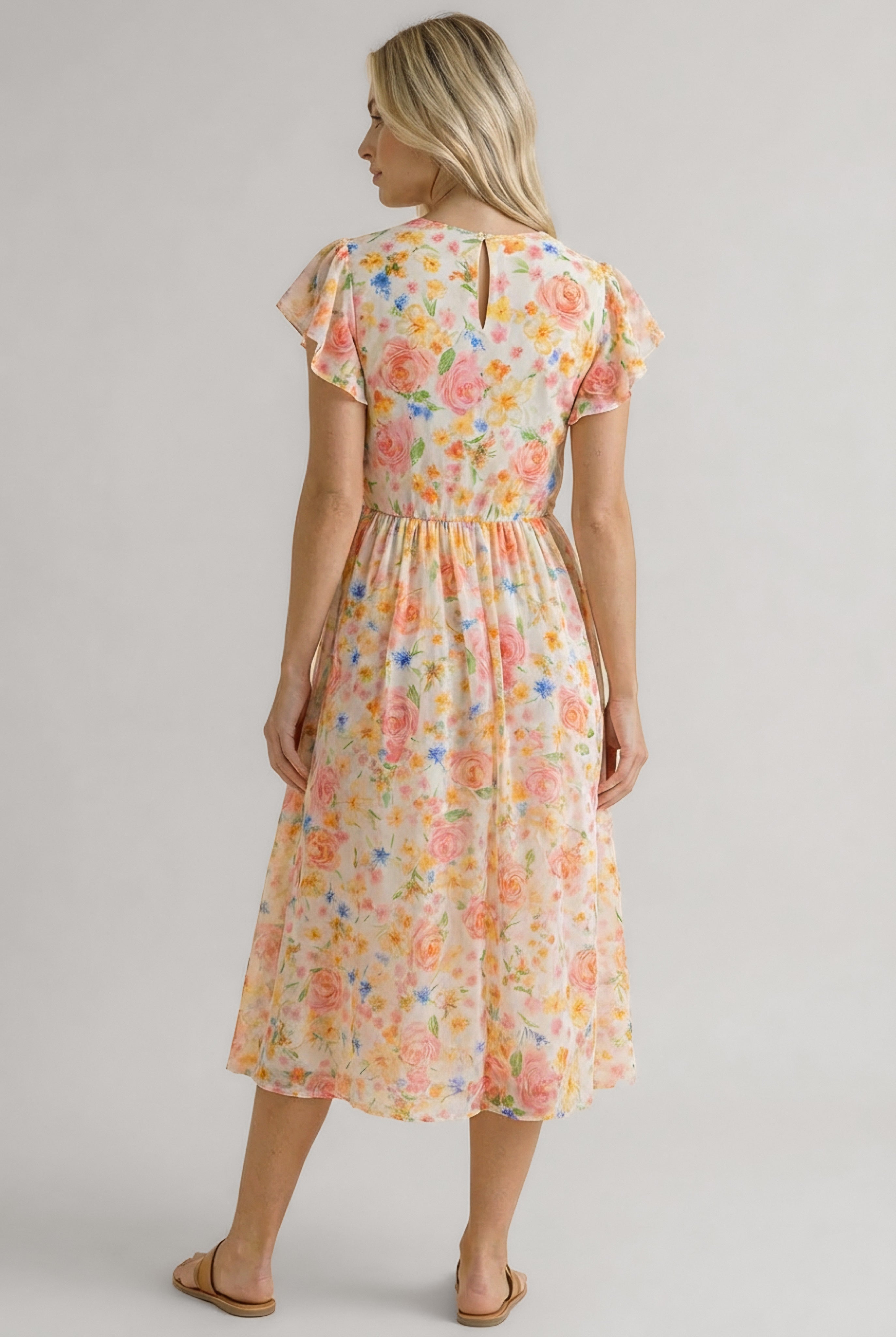 Woman with blonde hair wearing a midi floral dress with orange and pink flowers standing against a neutral background